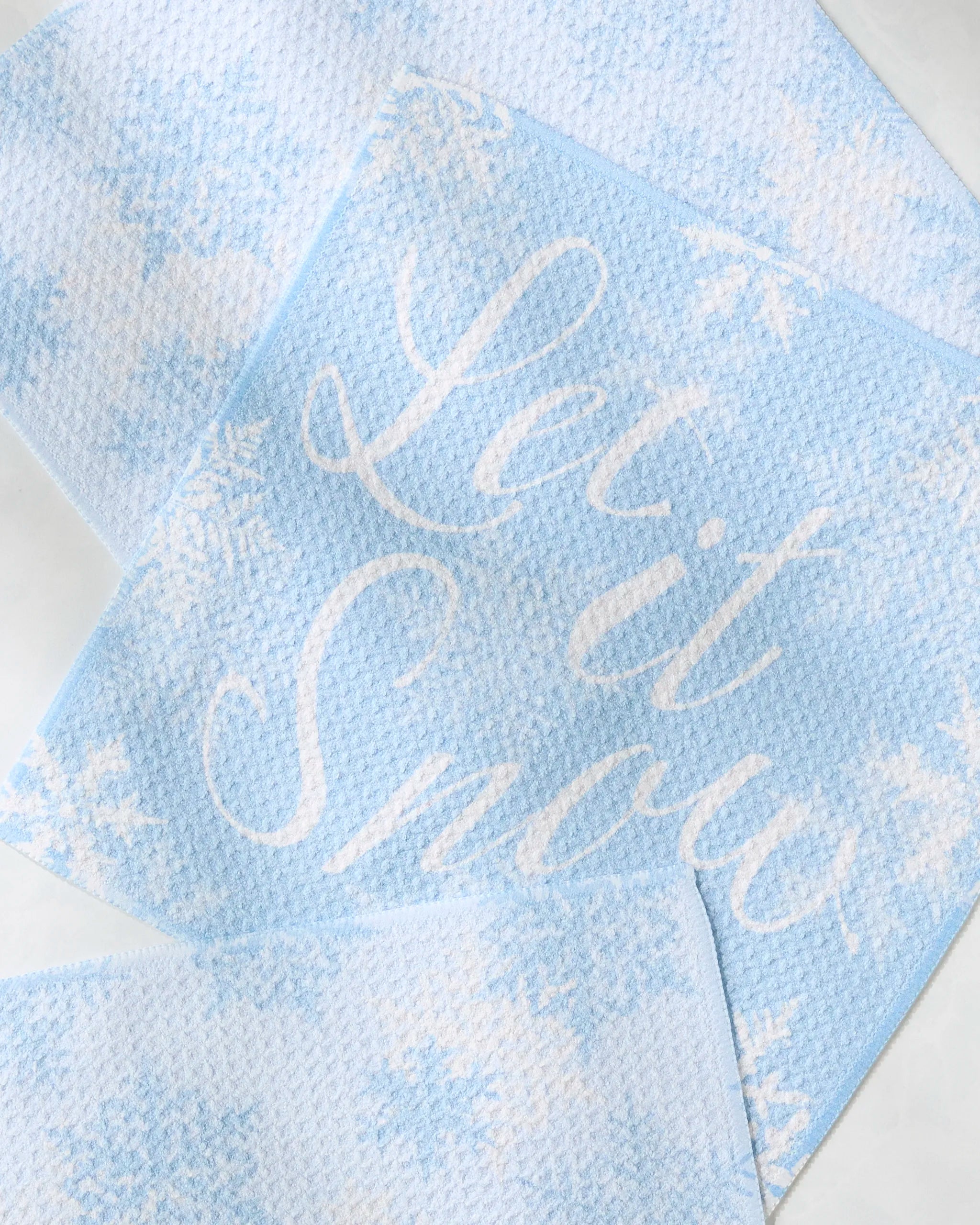 The Geometry Snow Frostin dishcloth features a light blue textured design with white snowflakes and "Let it Snow" in white script, perfect for adding a festive touch to your kitchen essentials.
