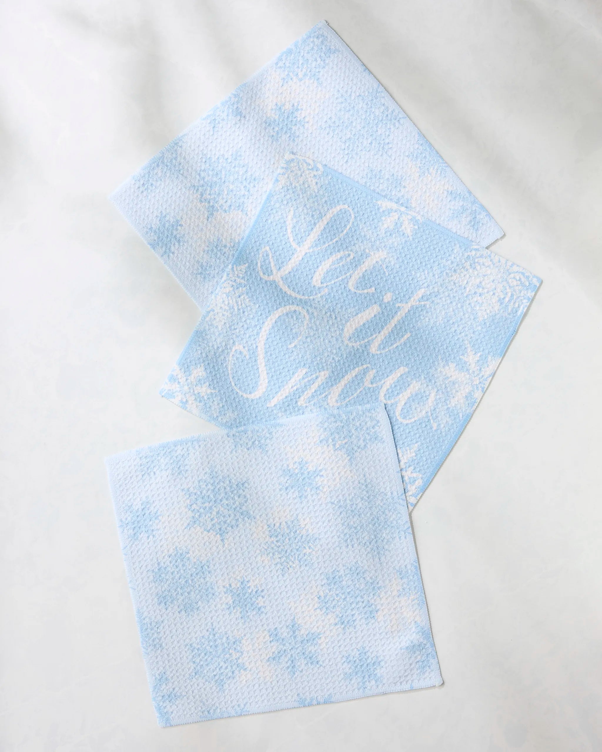The Geometry Snow Frostin set includes three blue absorbent dishcloths with white snowflake patterns—one featuring "Let it Snow" in white script—arranged on a light, textured surface.