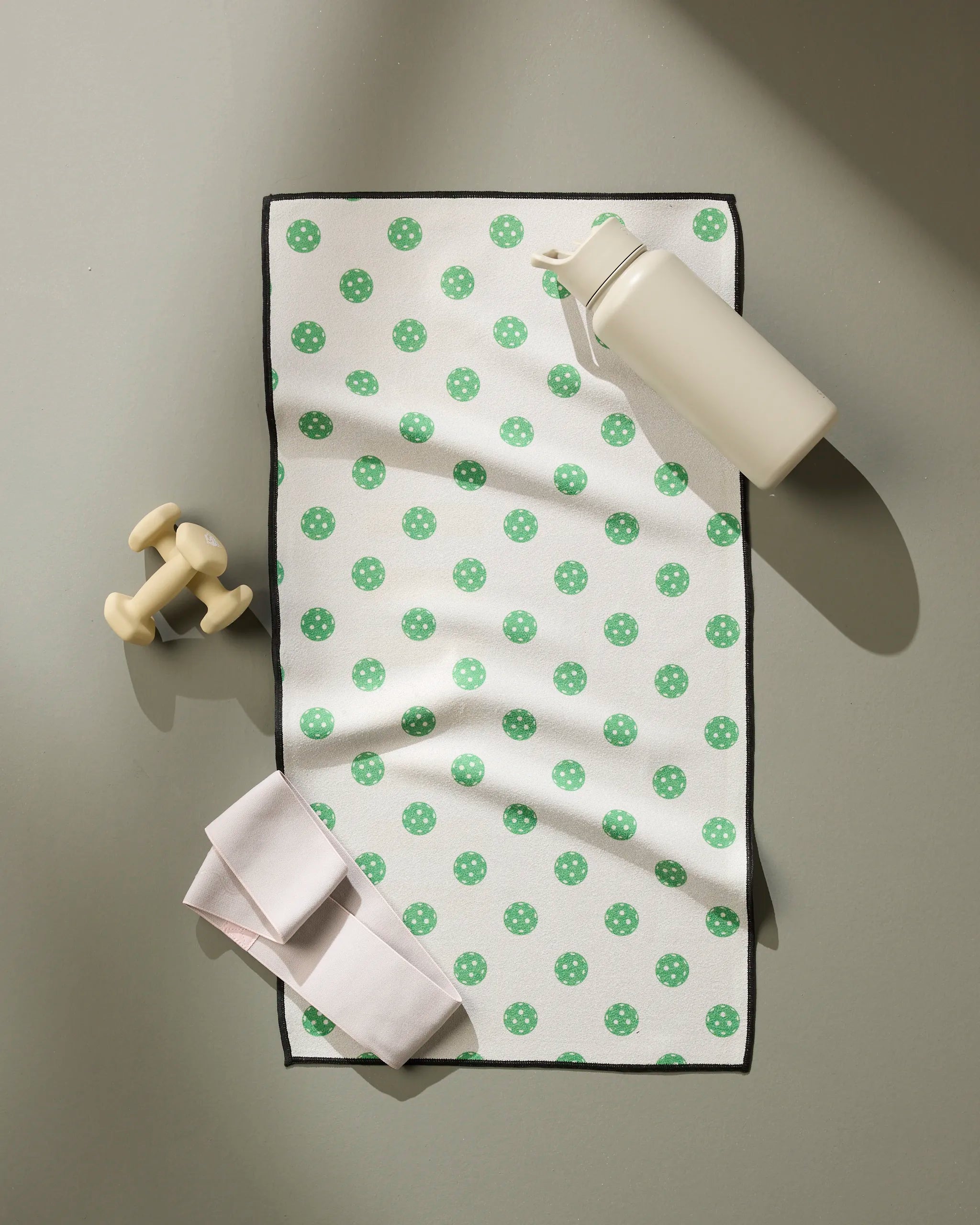 The Geometry Ace fitness towel in white with green polka dots is laid flat on a gray surface beside a white water bottle, light dumbbells, and a folded white band—perfect for your ultra-absorbent, fast-drying workout routine.