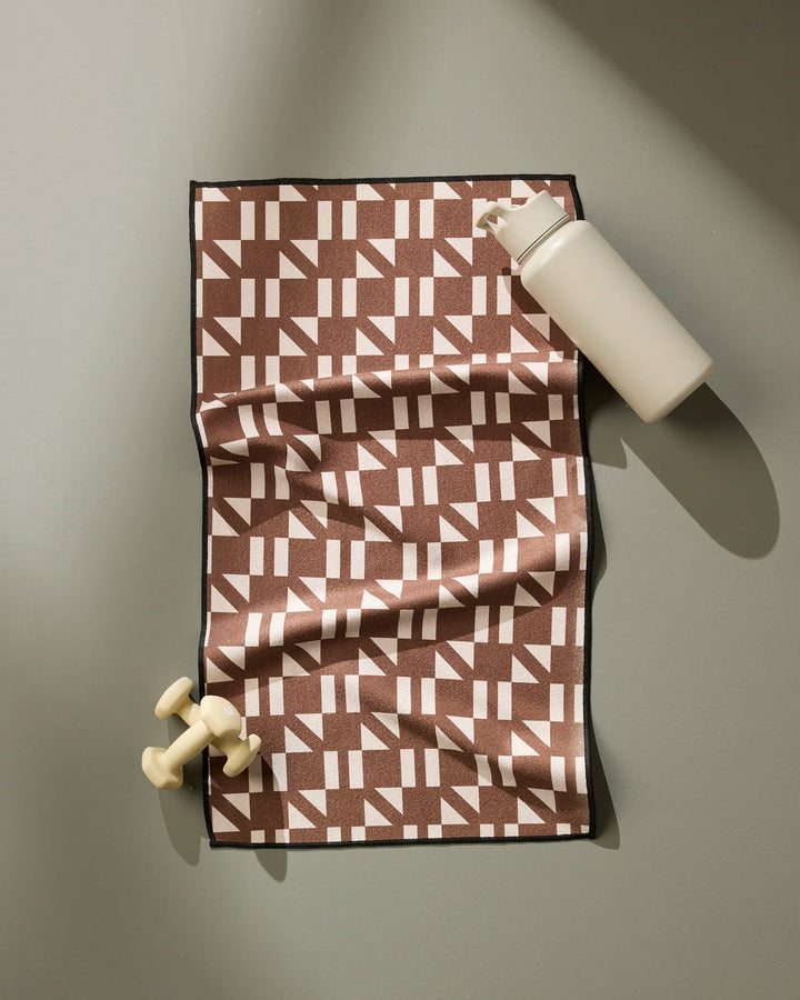 The Geometry Affogato fitness towel, featuring a patterned brown and white design, is displayed on a gray surface beside a light beige water bottle and small cream-colored dumbbells. Its ultra-absorbent fabric ensures fast drying during workouts.