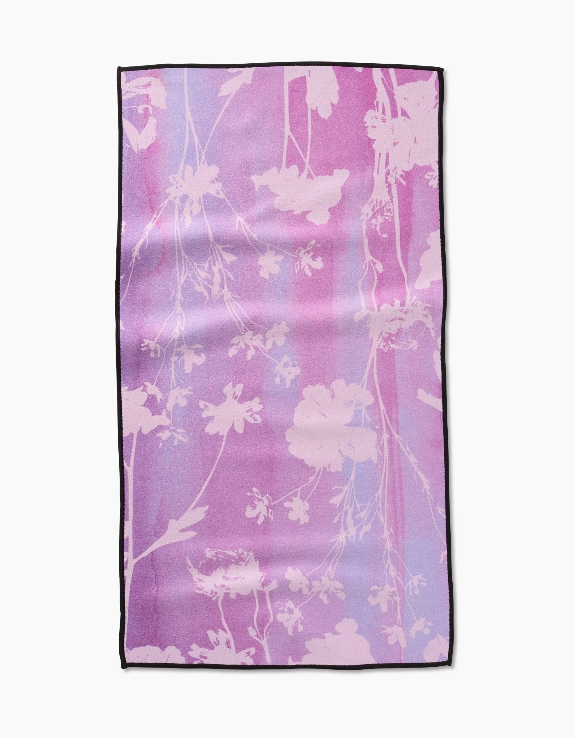 The GEOMETRY Bloom Blend is a rectangular fitness towel with a pink and purple gradient, white floral silhouettes, black edge piping, and made from ultra-absorbent recycled materials.