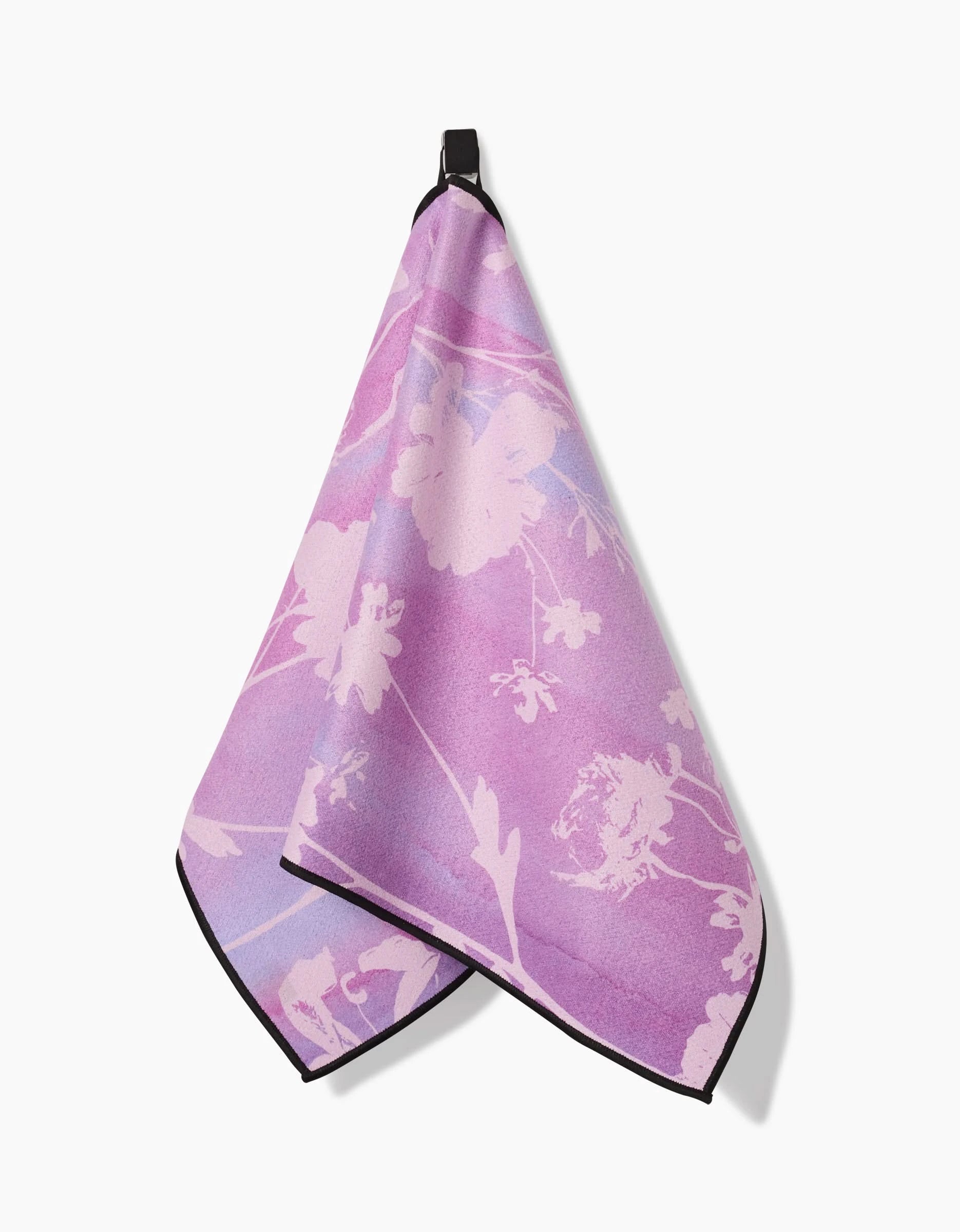 The GEOMETRY Bloom Blend fitness towel, crafted from recycled materials, features a pink and purple floral pattern with black trim and hangs neatly from a hook against a white background.