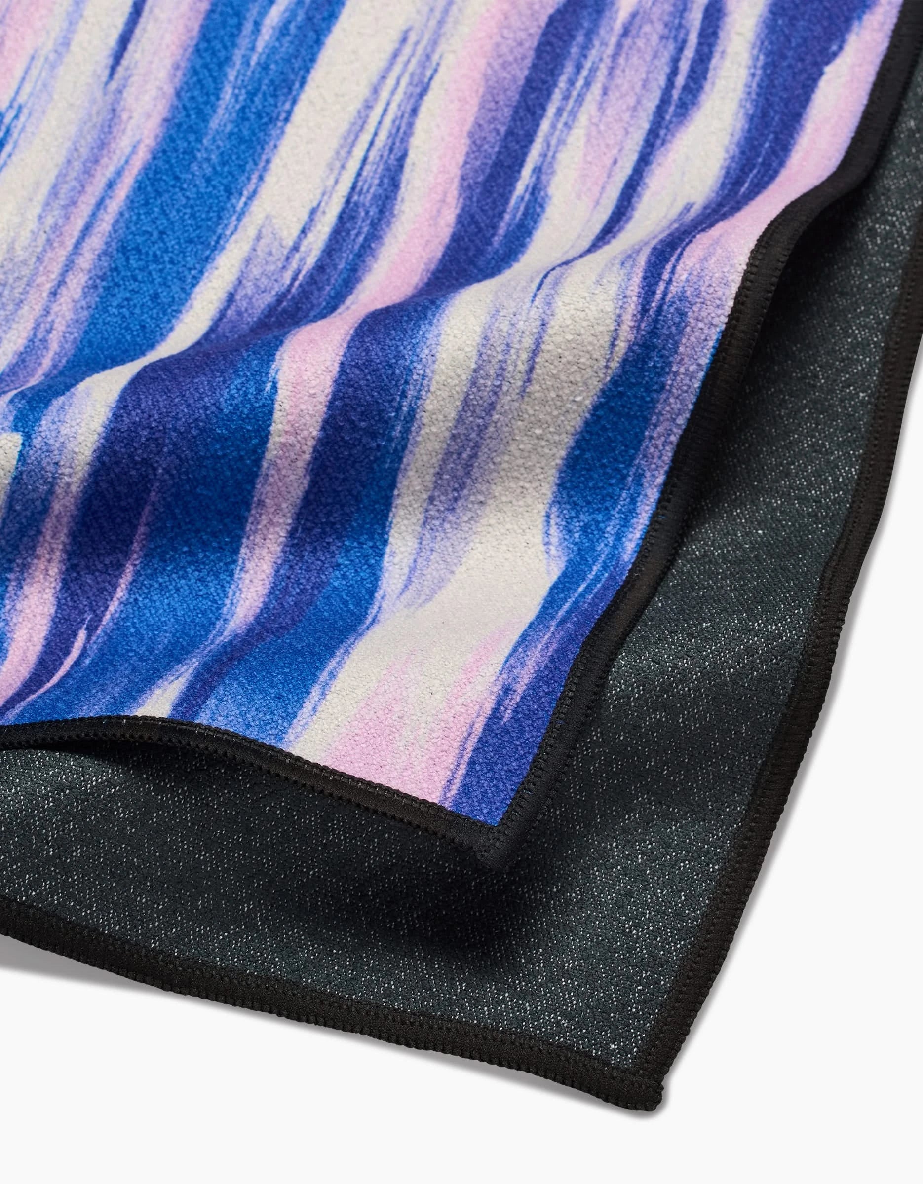 A close-up of two overlapping GEOMETRY Blue Blast fitness towels—one with a blue, purple, and pink abstract pattern and one in solid dark grey. Both are ultra-absorbent, made from recycled materials, and finished with black edging.