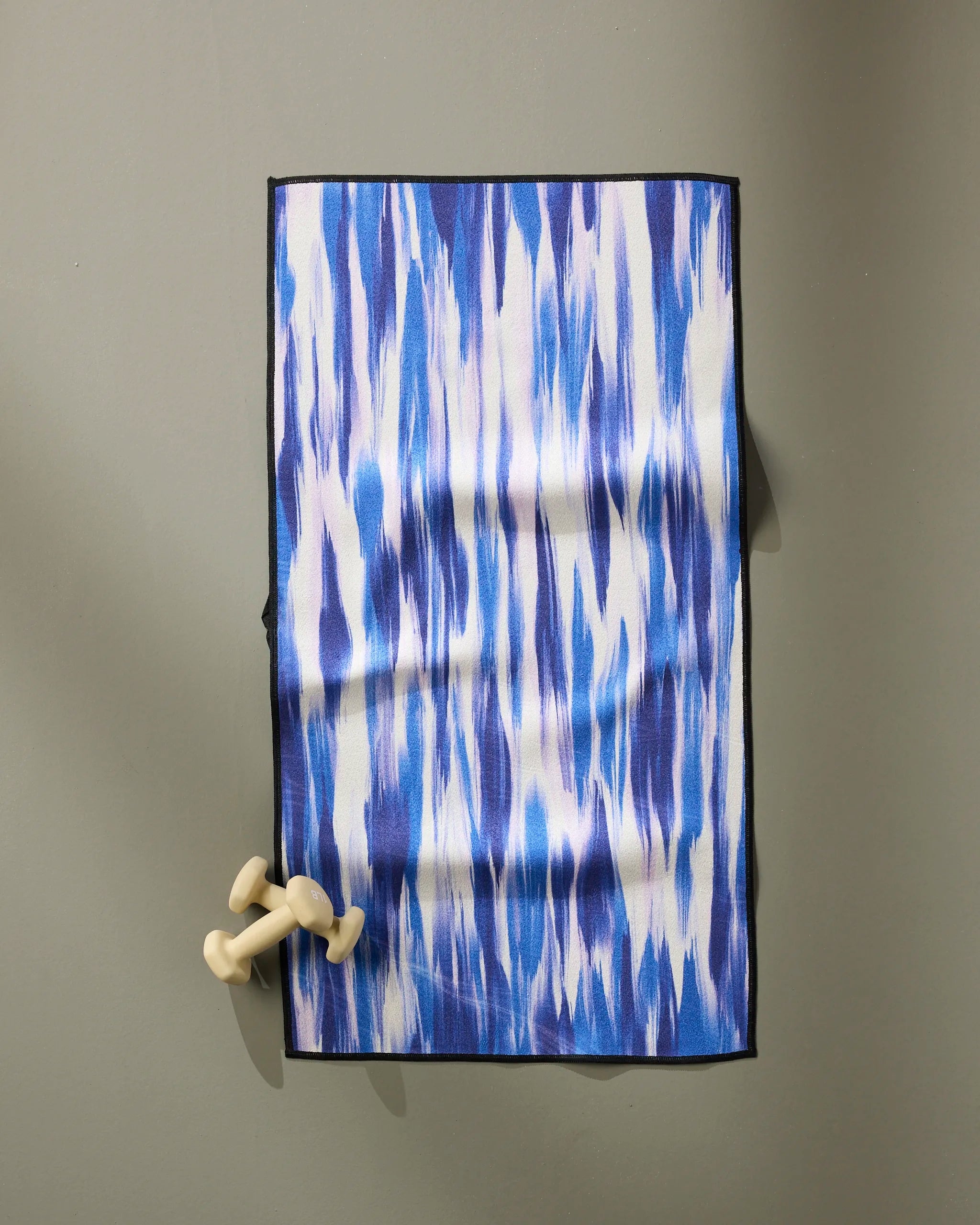 The GEOMETRY Blue Blast fitness towel, made from recycled materials and featuring a blue and white pattern, is spread on a gray surface with light-colored dumbbells placed on one corner.