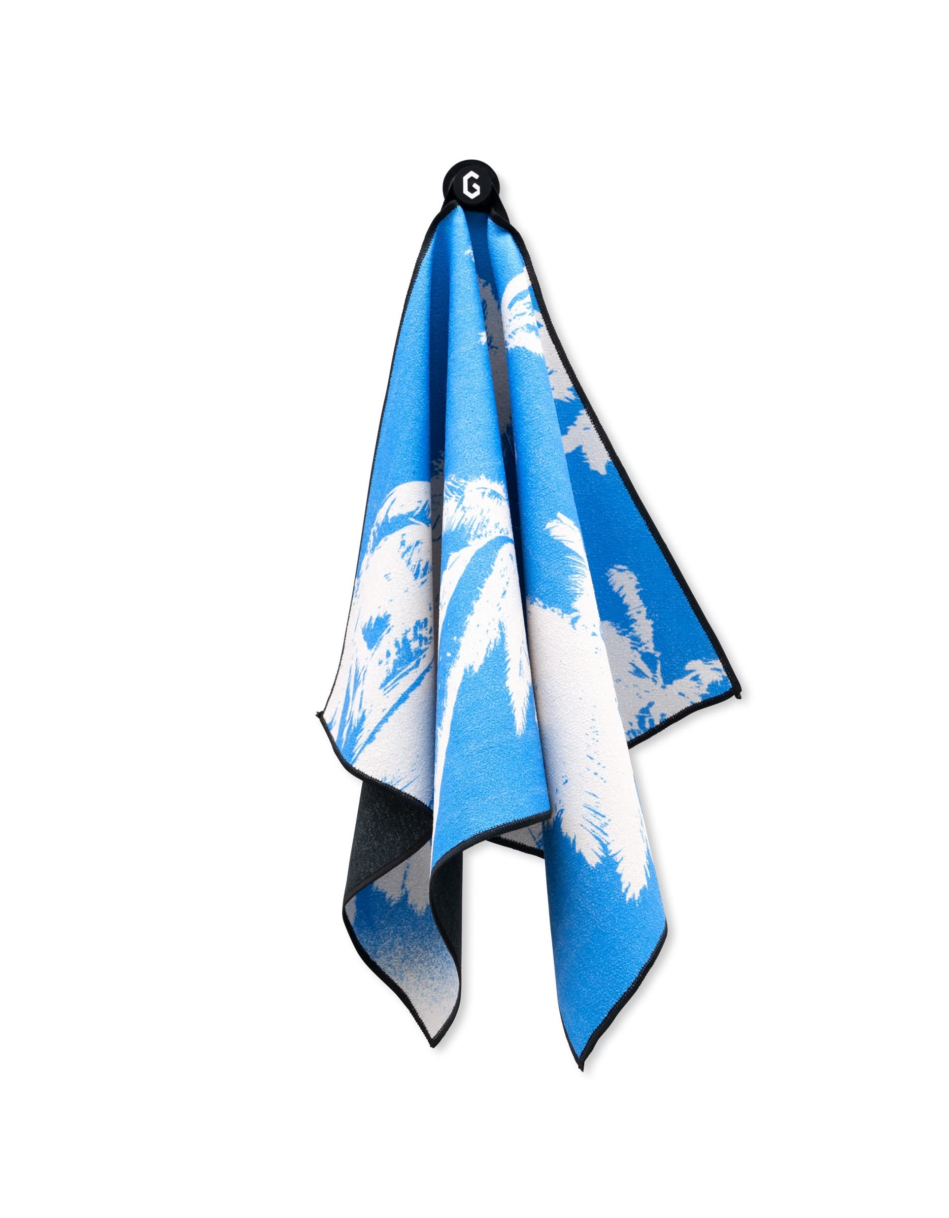 The GEOMETRY Magna - Blue Palms is an ultra-absorbent towel in blue and white with a palm tree pattern, hanging from a black loop featuring a white G.