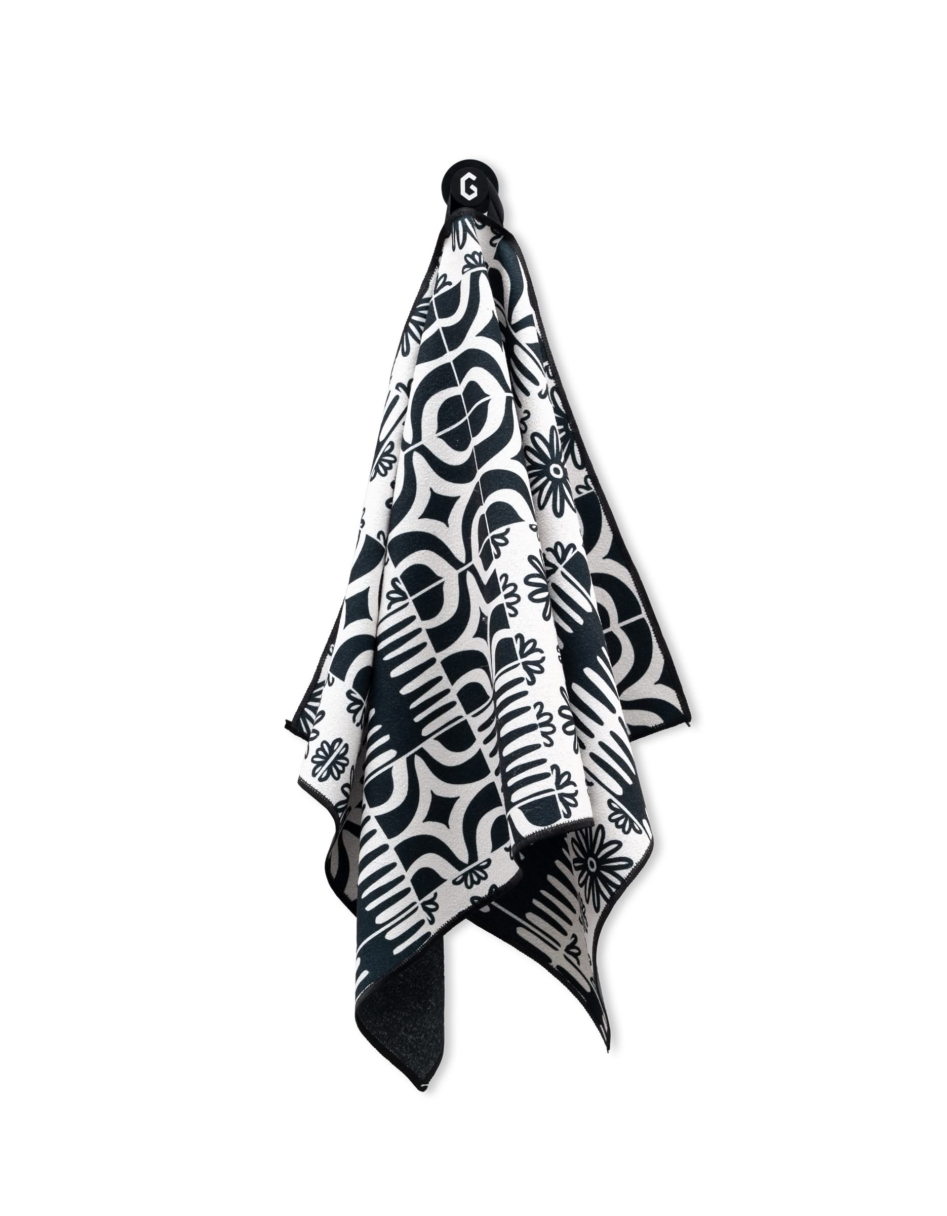 The GEOMETRY Magna - Bohemian Mosaic is a black and white ultra-absorbent gym towel featuring bold geometric and floral patterns, hanging by a black loop.