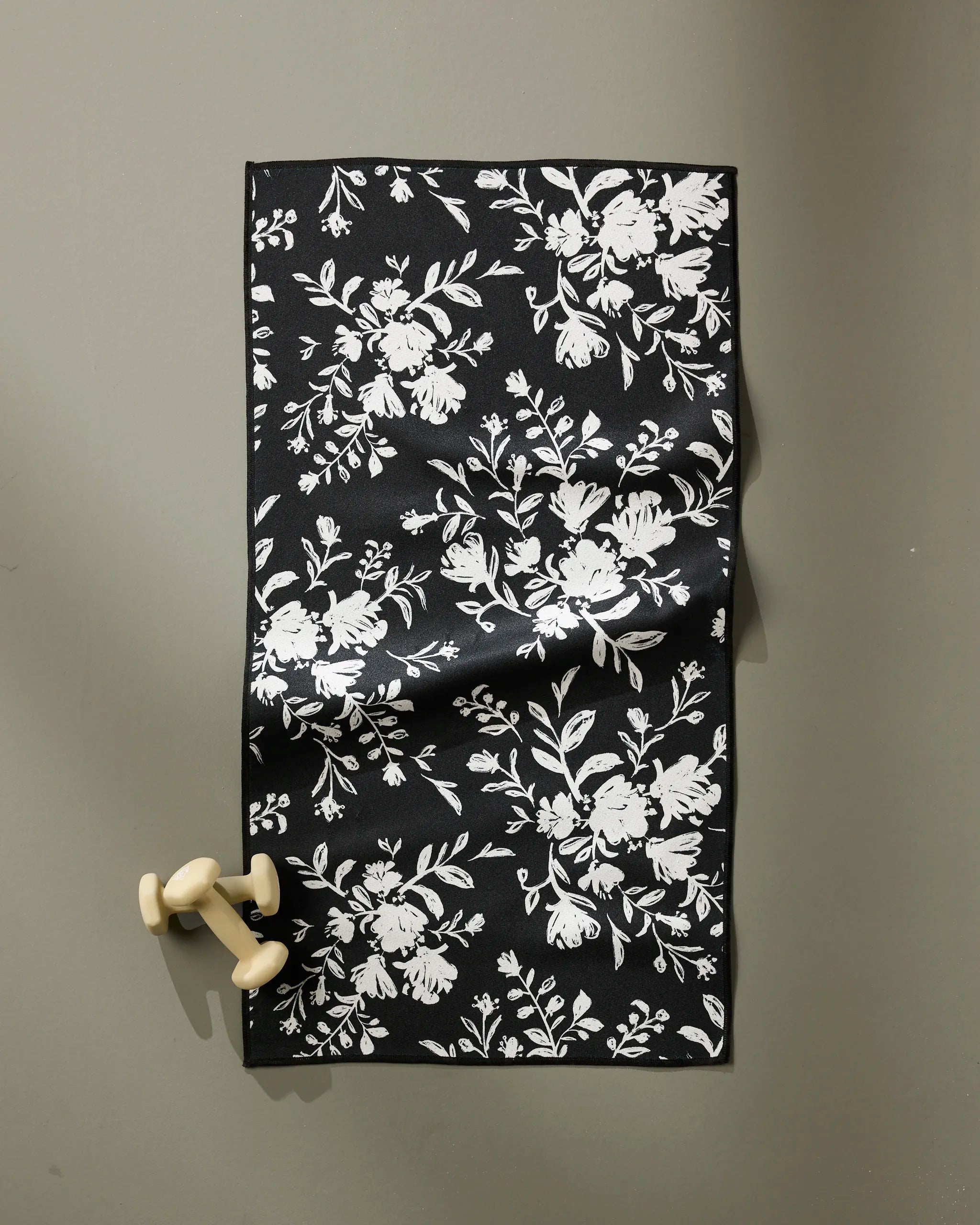 The Danelys Sidron Botanical Silhouettes Charcoal towel, featuring a white floral design, is laid on a gray surface with two small wooden dumbbells resting beside its ultra-absorbent fabric.