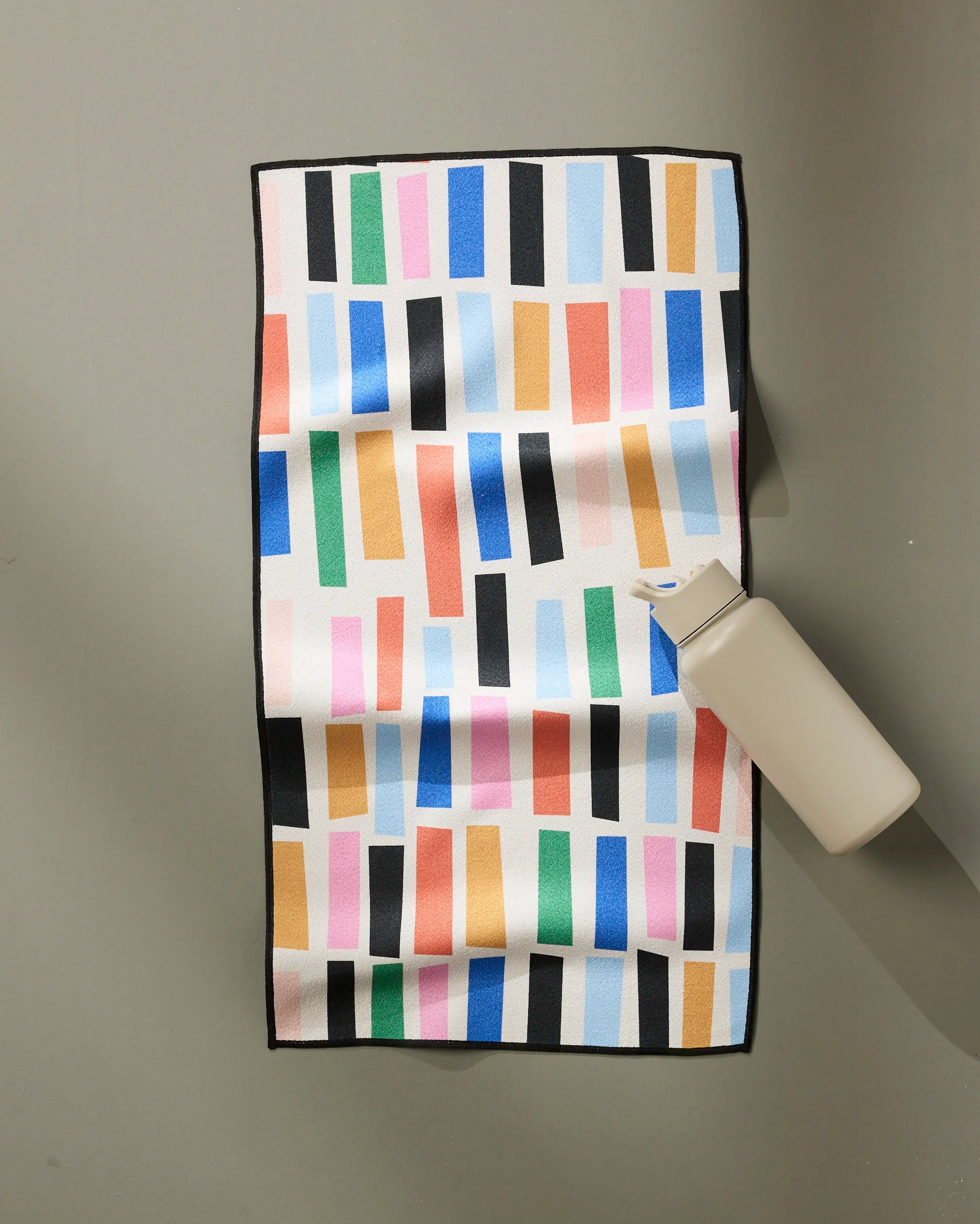 A beige water bottle sits on the Jennifer Bouron Bricks fitness towel, which is ultra-absorbent and features a light background with colorful vertical rectangles in blue, pink, green, orange, and black.