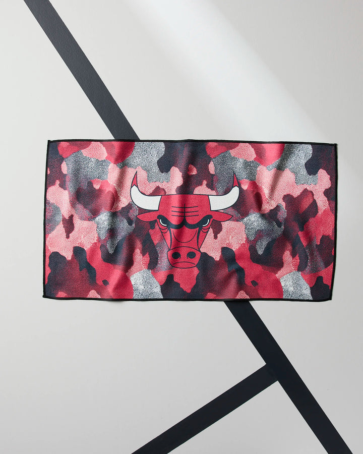 The NBALAB x Geometry Bulls Camo gym towel, featuring a red, black, white, and gray camouflage design with a stylized bulls head logo, hangs on a wall with two intersecting black diagonal lines.