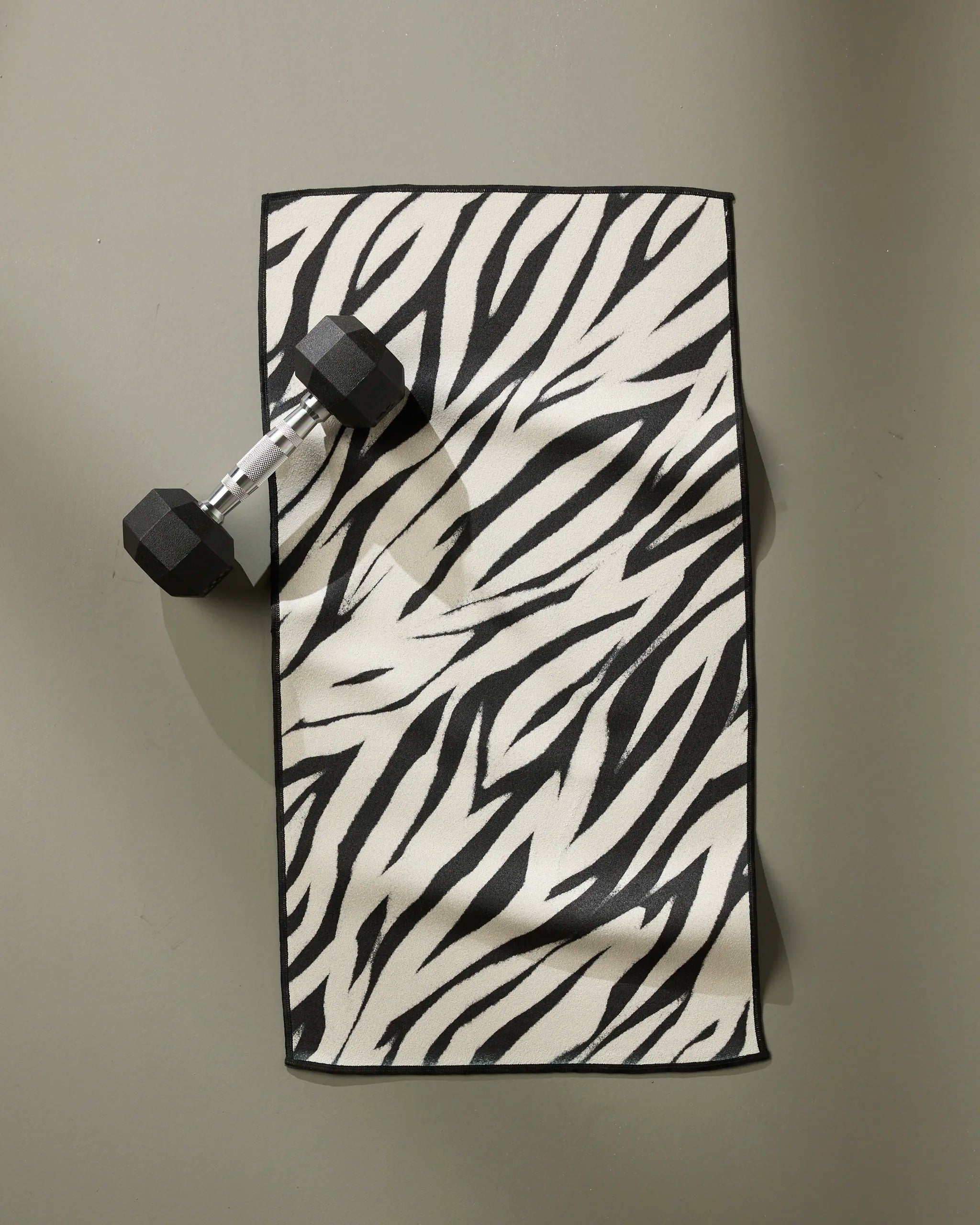 The GEOMETRY Chase fitness towel, featuring ultra-absorbent recycled materials and a black-and-white zebra stripe design, is displayed on a gray surface with a black dumbbell placed on its upper left corner.