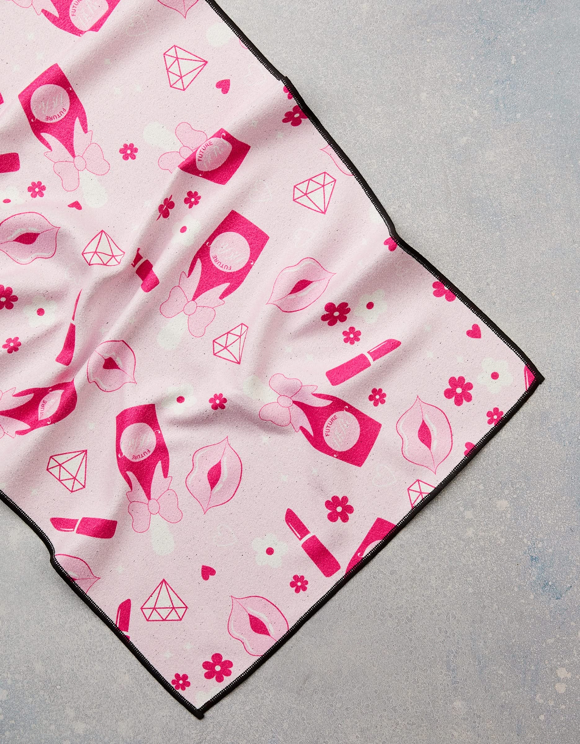 The GEOMETRY Cheers fitness towel, made from recycled materials, showcases lipstick, lips, flowers, diamonds, and compacts on a light pink background with black edges, displayed flat on a light gray textured surface.