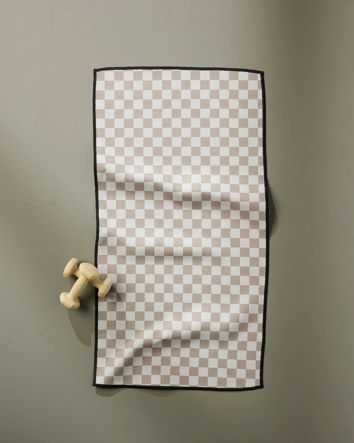The GEOMETRY Chic Checker fitness towel, made from recycled materials in a beige and white checkered design, is laid flat on a gray surface with a small cream dumbbell near its left edge.
