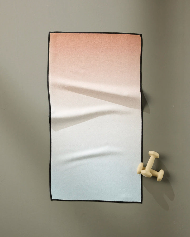 A GEOMETRY Core fitness towel with a peach-to-white gradient, made from recycled materials, lies flat on a gray surface. Ultra-absorbent, it features small cream dumbbells near the bottom right corner.