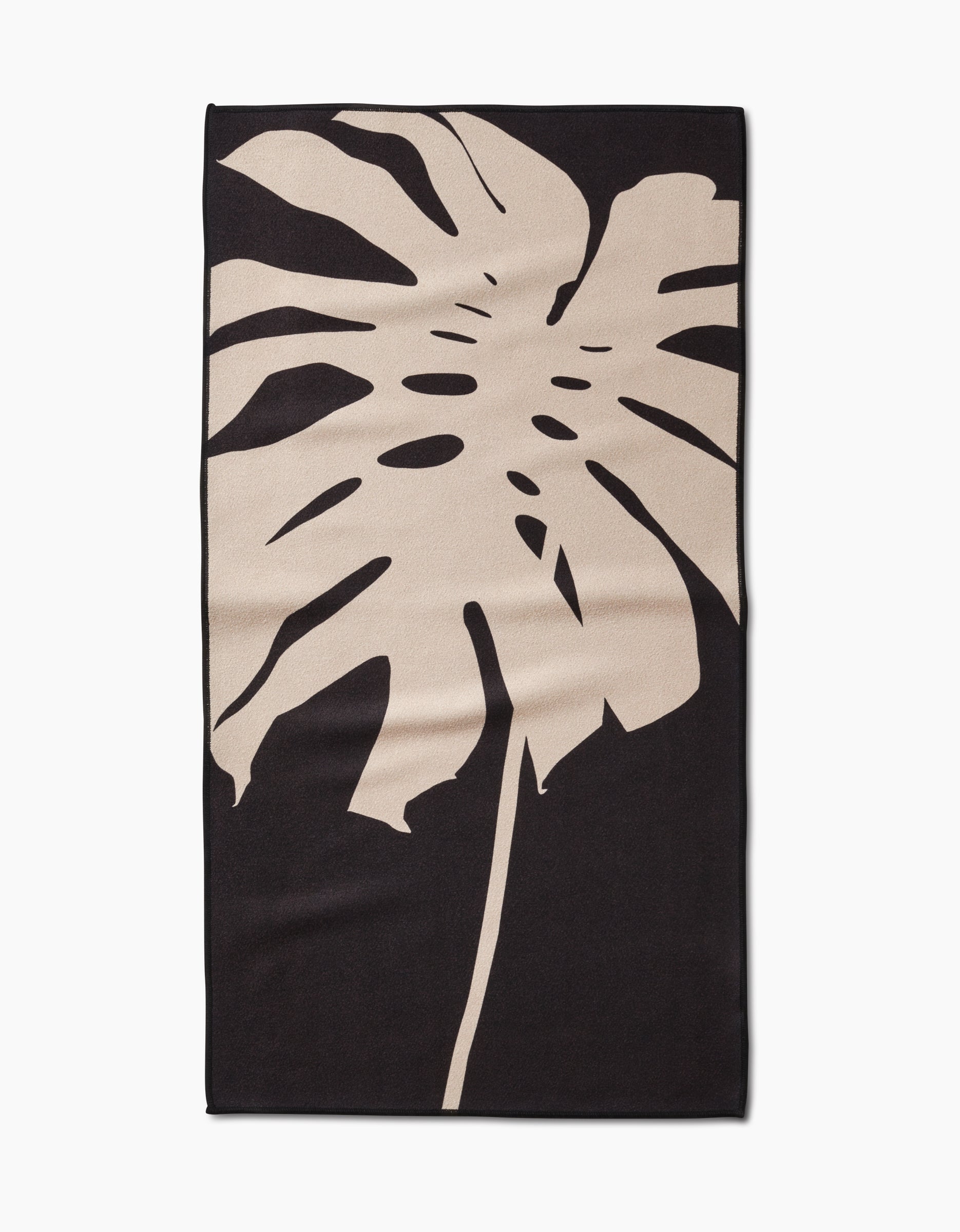 The PrintedPlant Cream Monstera Fitness Towel is rectangular, made from ultra-absorbent recycled materials, and features a large light beige monstera leaf design on a solid black background.