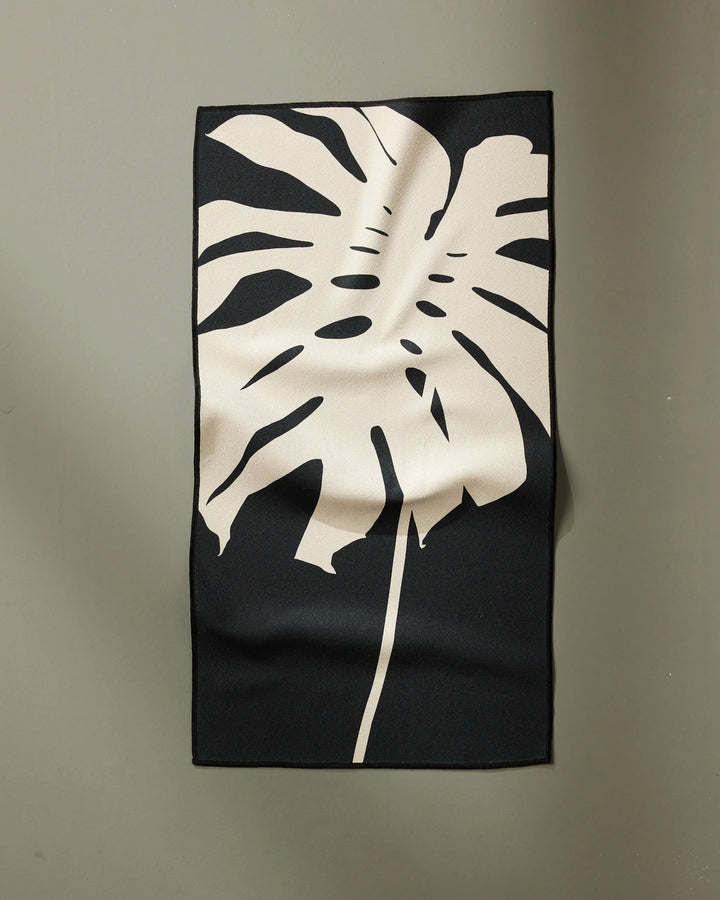 The PrintedPlant Cream Monstera is a rectangular black fitness towel made from recycled materials, featuring a large beige monstera leaf design and ultra-absorbent fabric with gentle folds, shown hanging on a plain light gray wall.
