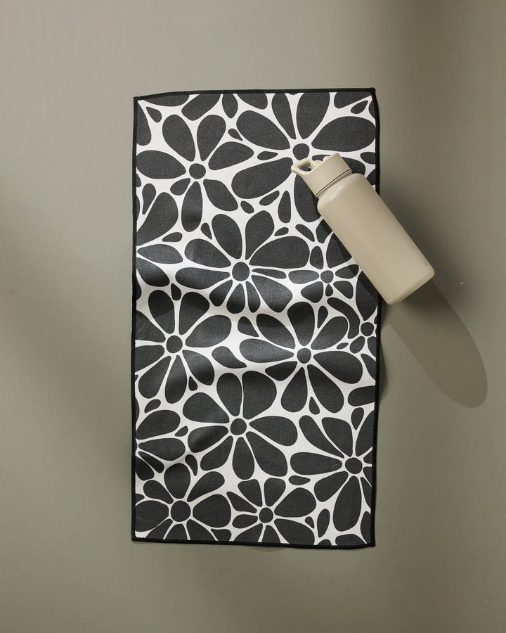A beige water bottle sits on the GEOMETRY Cute Flowers towel, featuring a bold black and white floral pattern, on a smooth light gray surface.