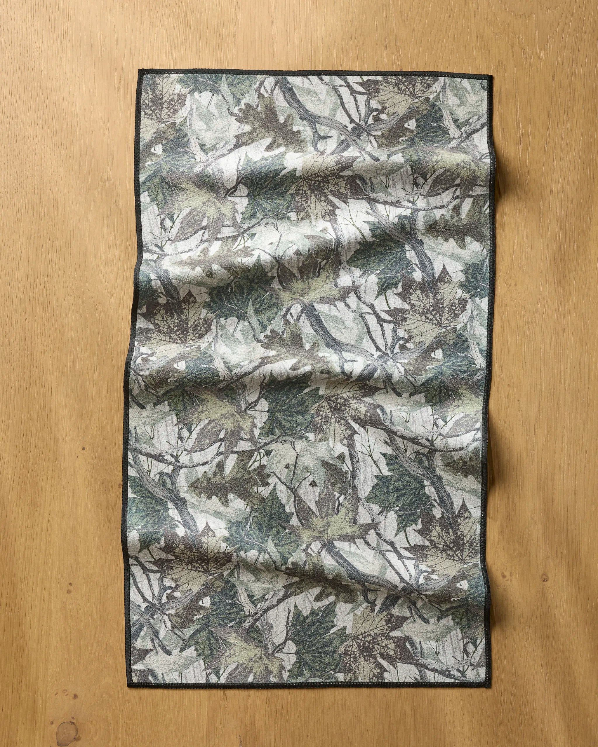The Geometry Dad Camo rectangular fitness towel, featuring a green and gray leafy pattern with a sleek black border, is fast-drying and ultra-absorbent. It’s shown laid out on a light wooden surface, displaying its stylish wrinkle look.