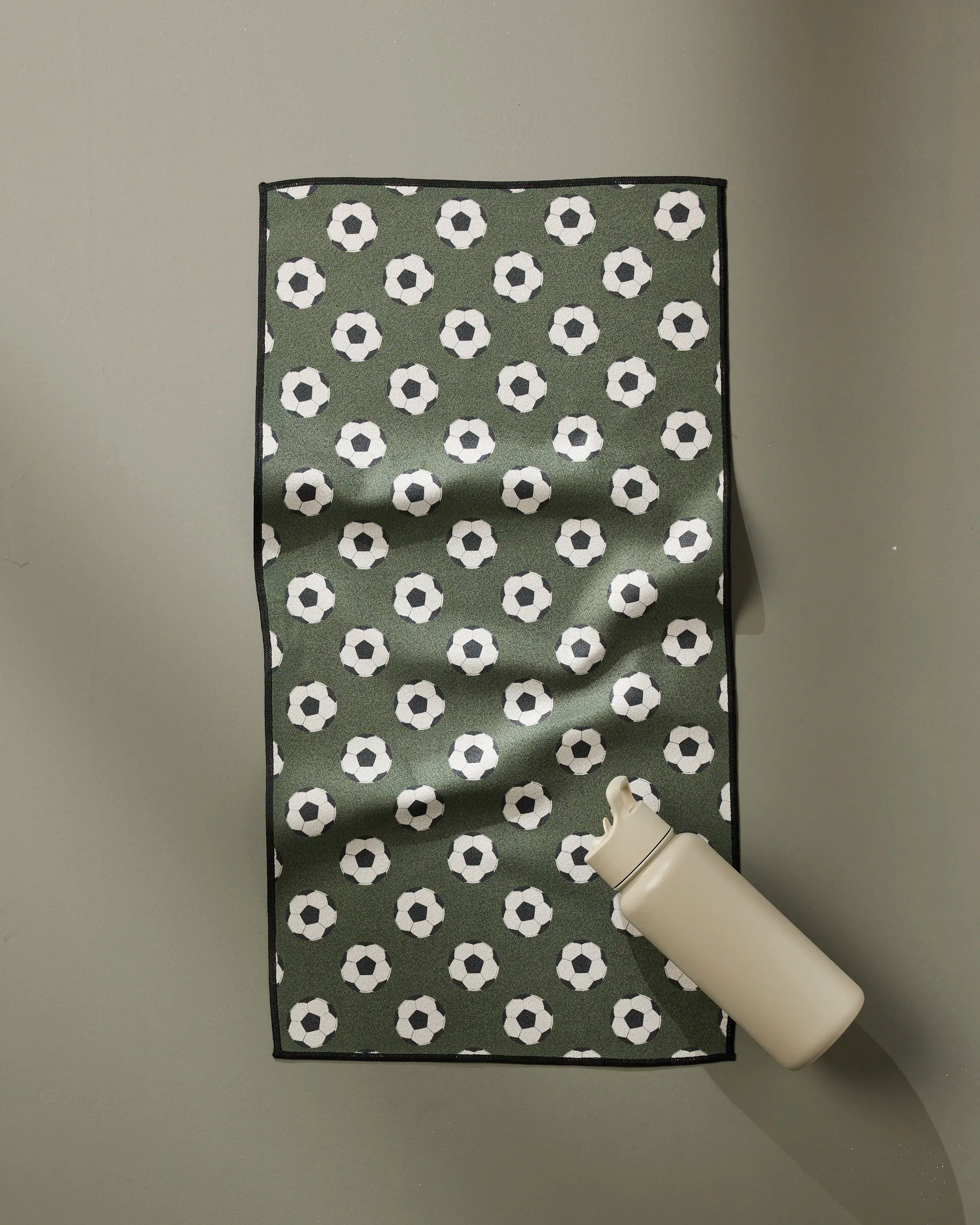 The Geometry Dribble towel, featuring a repeating soccer ball pattern in white and black, is laid flat on a pale green surface with a beige water bottle positioned in the lower right corner.