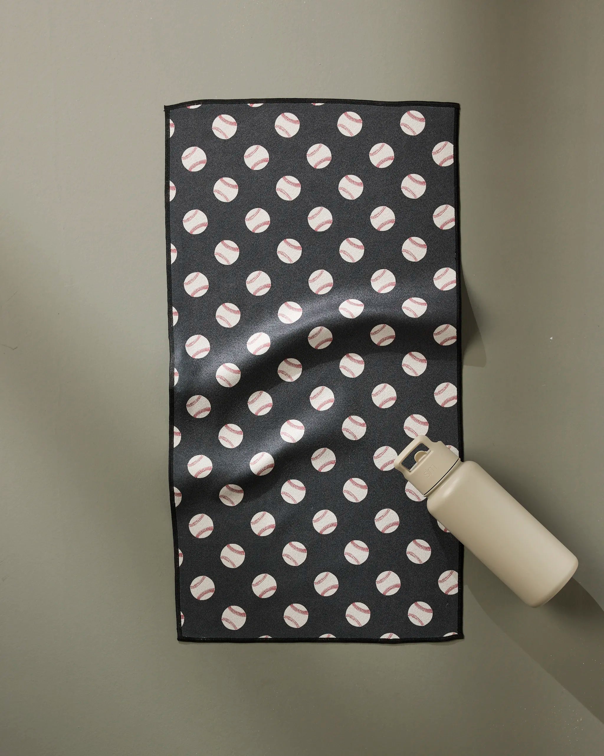 The Geometry Fastball towel, featuring a black design with a repeating baseball pattern, lies flat on a light gray surface with a beige water bottle at its lower right corner. Ultra-absorbent and fast drying, it's ideal for workouts or daily use.