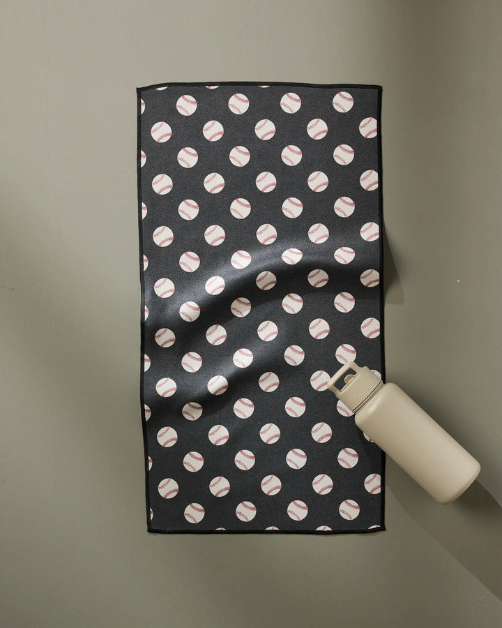 The Geometry Fastball towel, featuring a black design with a repeating baseball pattern, lies flat on a light gray surface with a beige water bottle at its lower right corner. Ultra-absorbent and fast drying, it's ideal for workouts or daily use.