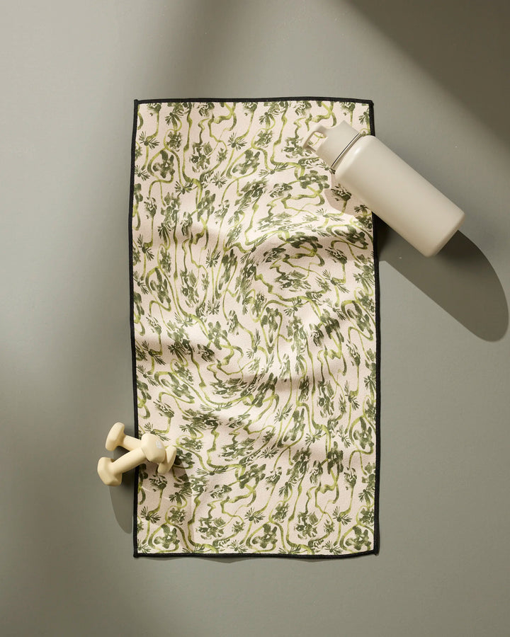 The Floral Swirls Green fitness towel by Geometry, made from recycled materials, is spread on a gray surface with cream dumbbells and a gray water bottle on top. Diagonal shadows accent the green and white patterned towel.