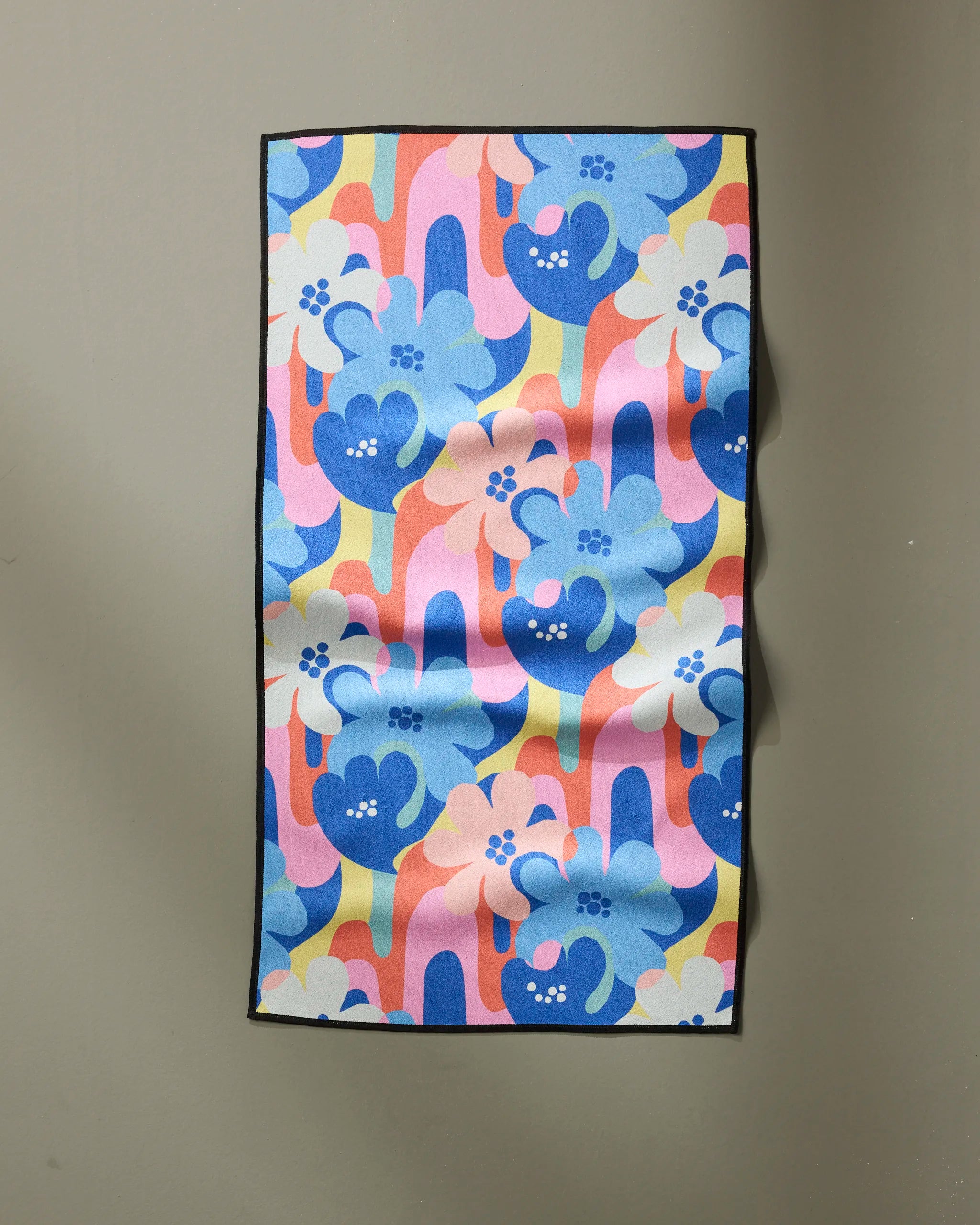 The "Flower Powers" fitness towel by Masha Volnova features a colorful abstract floral print in blue, pink, yellow, and orange. Made from recycled materials with a black border, it hangs on a neutral wall showing visible fabric creases.