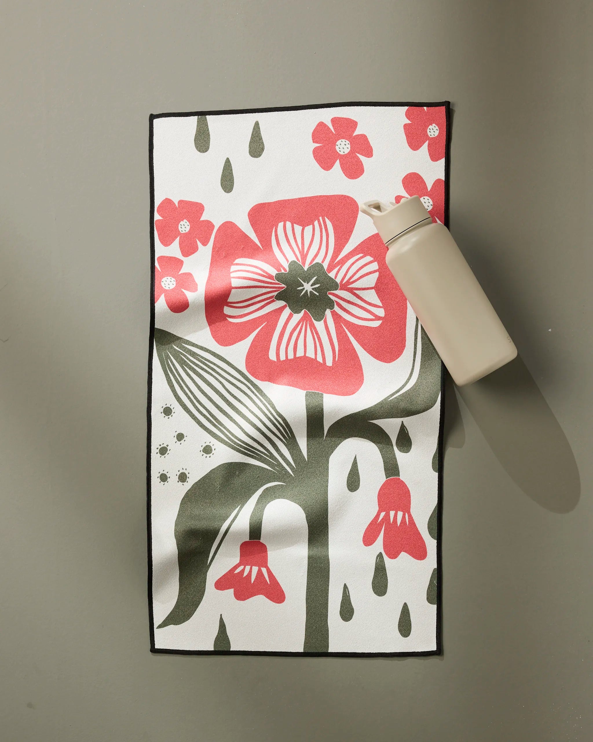 The Ash Cascade Flower Stamp fitness towel, ultra-absorbent and fast drying, features a large red and green floral design with smaller accents on white. It’s shown flat on gray beside a beige water bottle in the upper right corner.