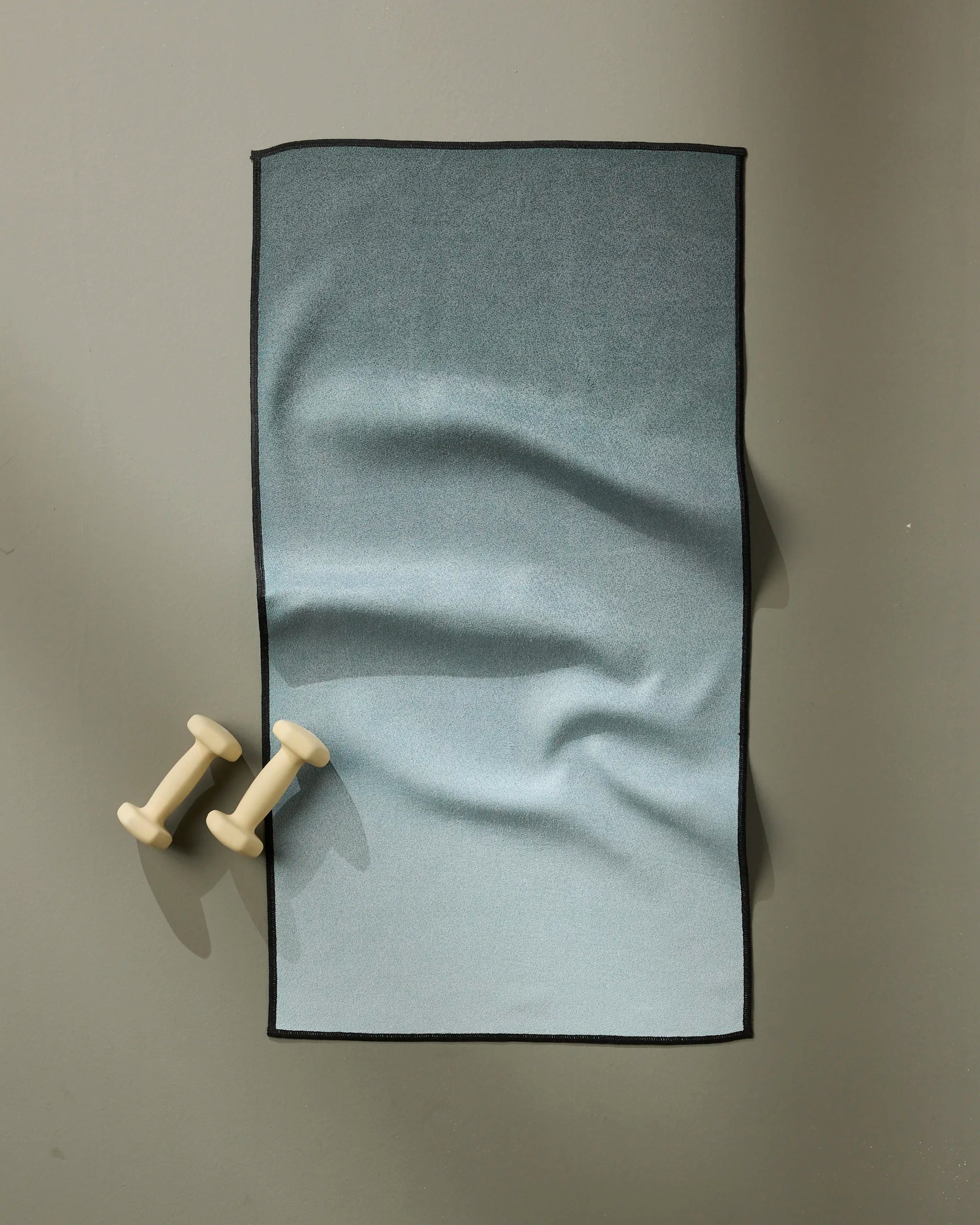 A pair of small, light-colored dumbbells sit on a gray surface next to a neatly folded GEOMETRY Foundation ultra-absorbent blue gradient fitness towel with black edges.