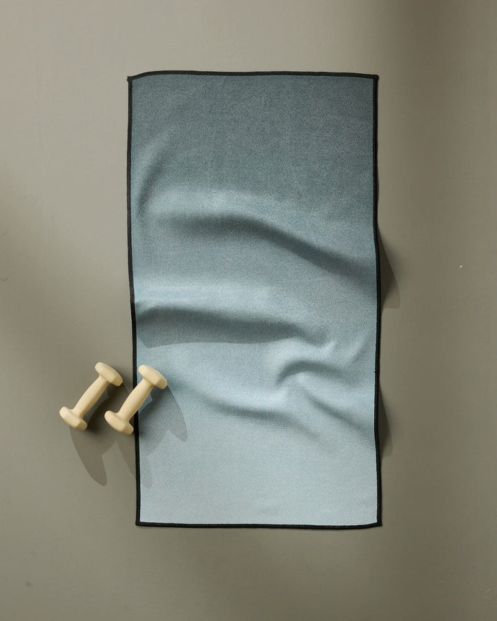 A pair of small, light-colored dumbbells sit on a gray surface next to a neatly folded GEOMETRY Foundation ultra-absorbent blue gradient fitness towel with black edges.