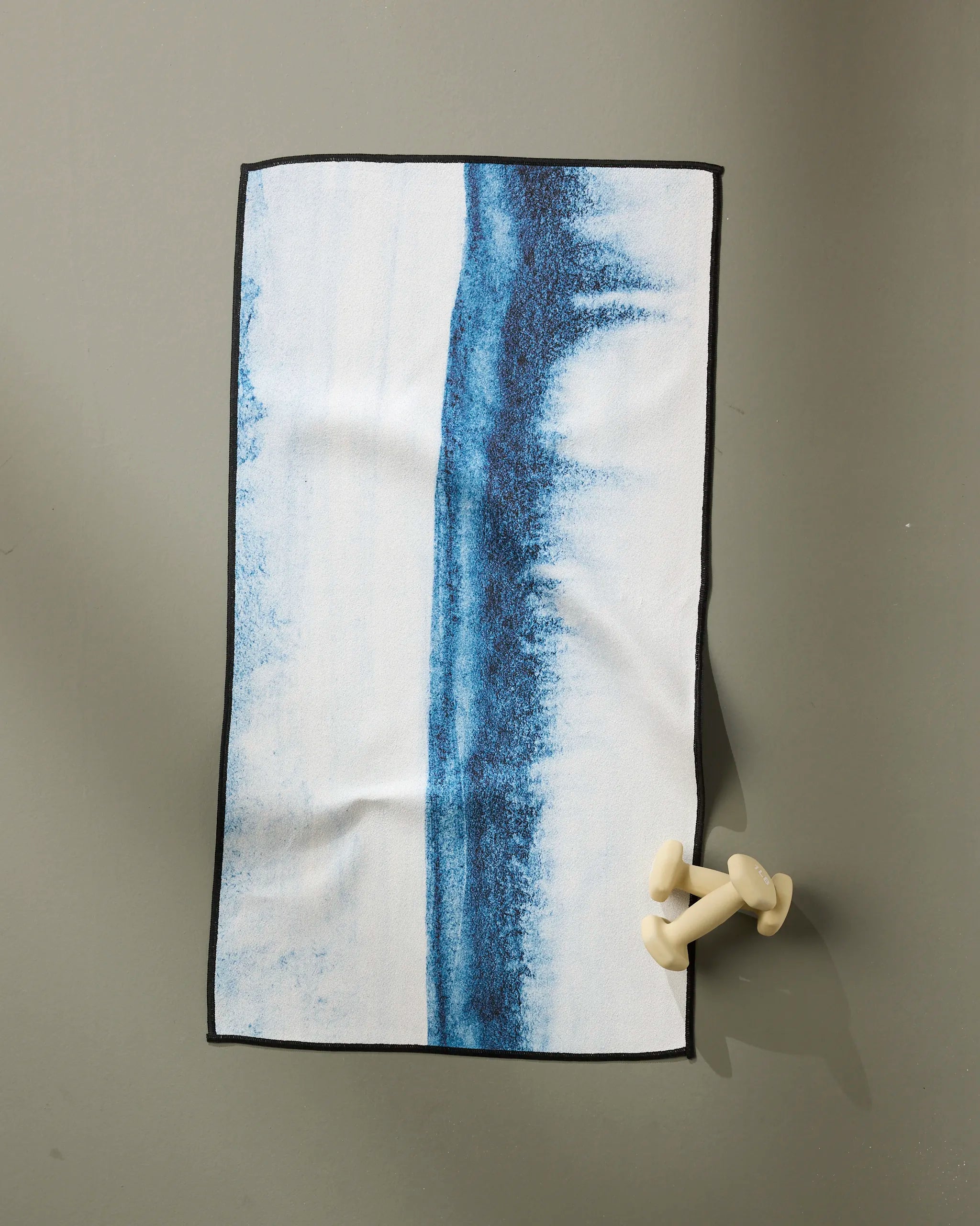 The GEOMETRY "In The Distance" rectangular fitness towel features a blue, wave-like center stripe and is ultra-absorbent. It’s displayed on a gray surface with two small white dumbbells near one corner.