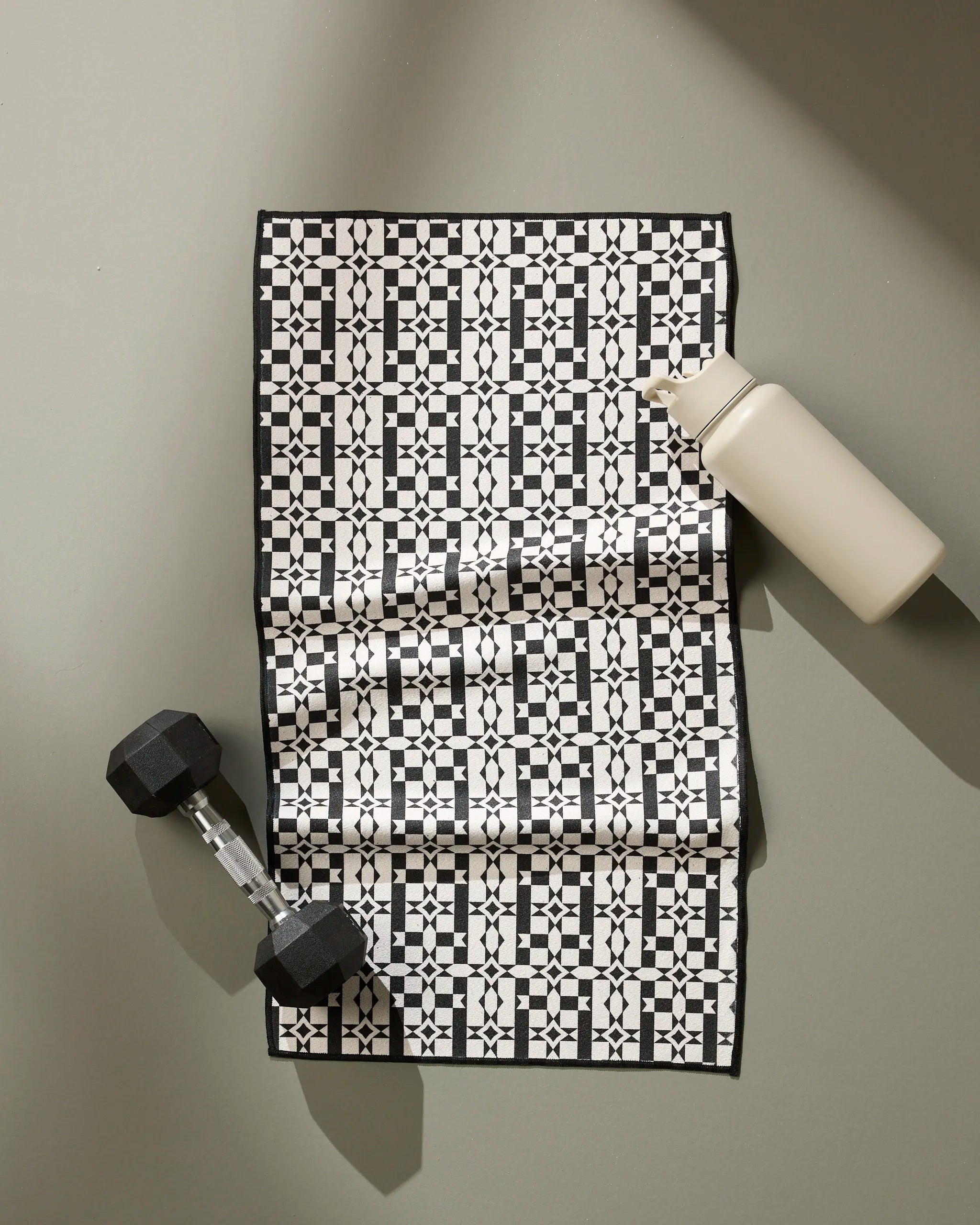 The Geometry Intermission ultra-absorbent fitness towel, featuring a black-and-white pattern, lies on a gray surface with a black dumbbell in the left corner and a white water bottle diagonally placed in the top right corner.