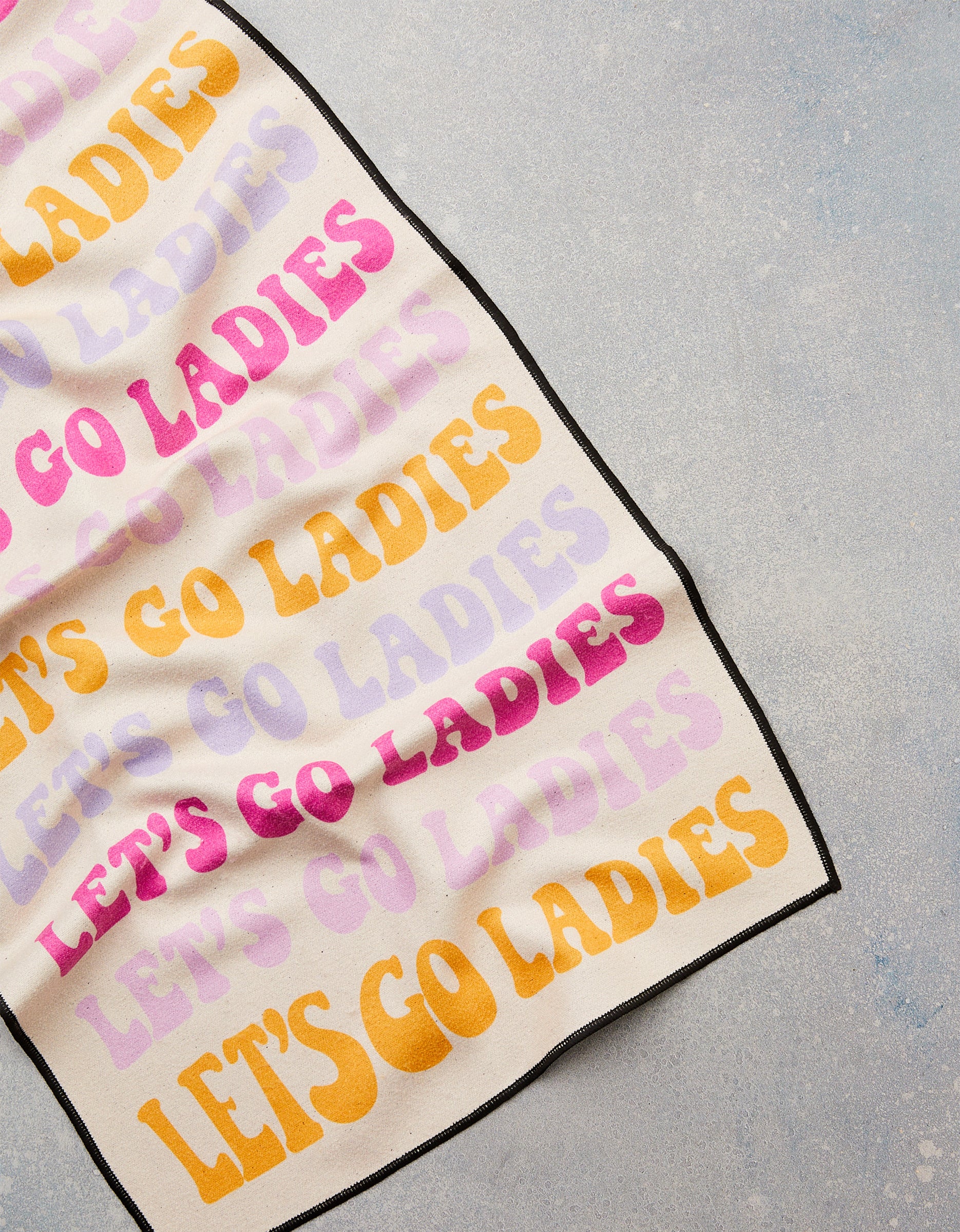 The GEOMETRY “Ladies Only” Fitness Towel, made from recycled materials, features bold yellow, pink, and lavender “LET’S GO LADIES” text on a light gray textured surface.