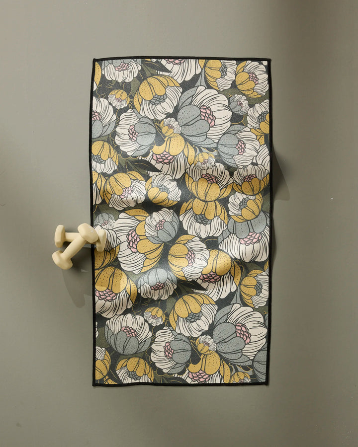 The Ash Cascade Lakeside Bloom towel, featuring white, yellow, and grey stylized floral patterns, hangs on a cream double hook against a plain grey background.