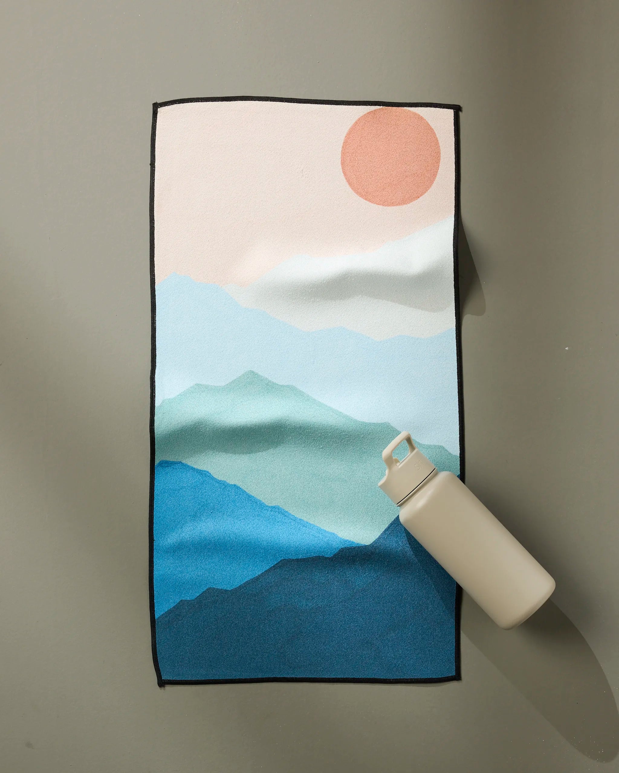 A beige water bottle sits on the GEOMETRY Fitness towel, "Landscape," which features blue mountains, a soft pink sun, and a pale sky on a neutral surface.