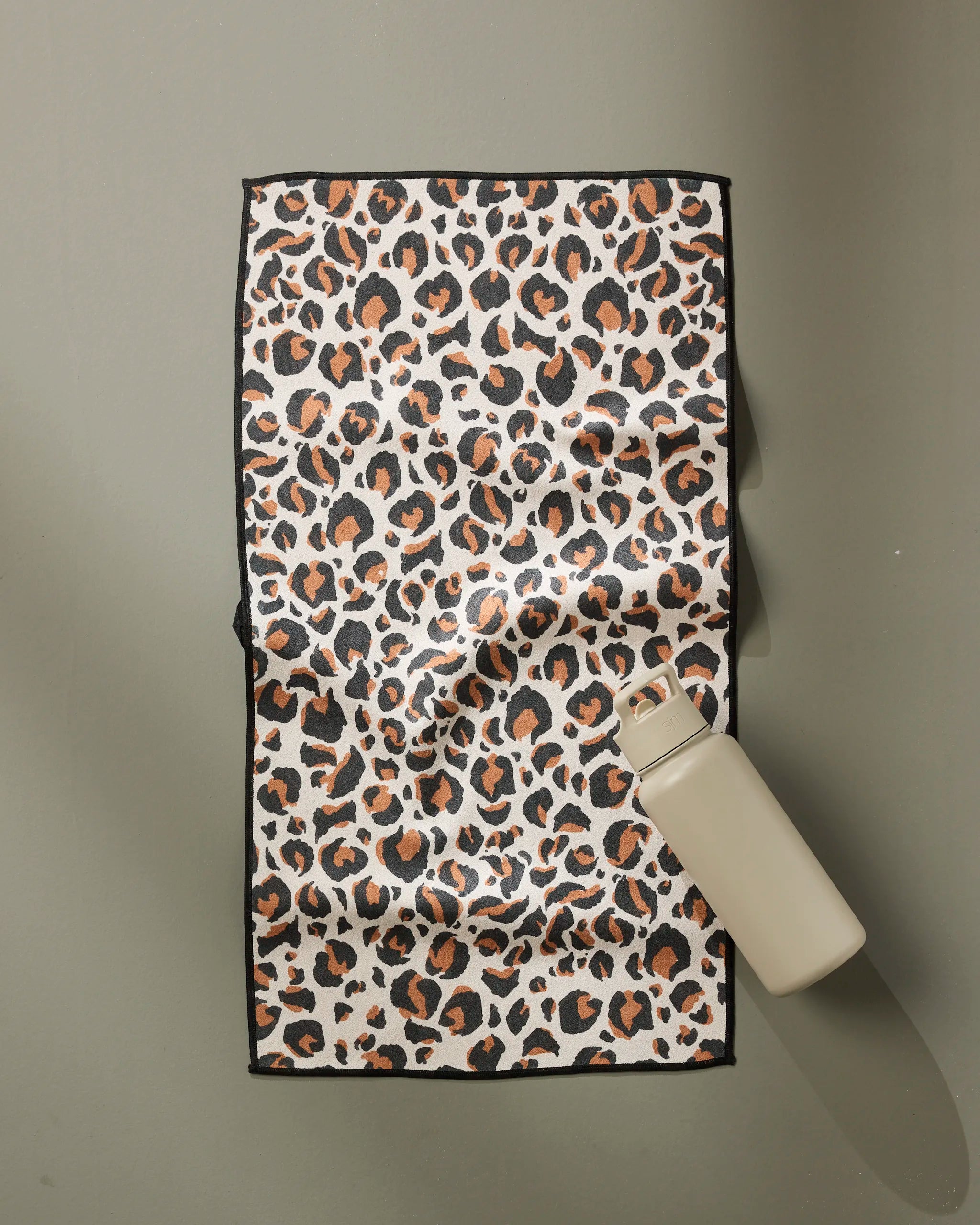 A beige water bottle sits on the GEOMETRY Leopard fitness towel, which features an ultra-absorbent design and a black, brown, and tan leopard print pattern, set against a plain light gray surface.