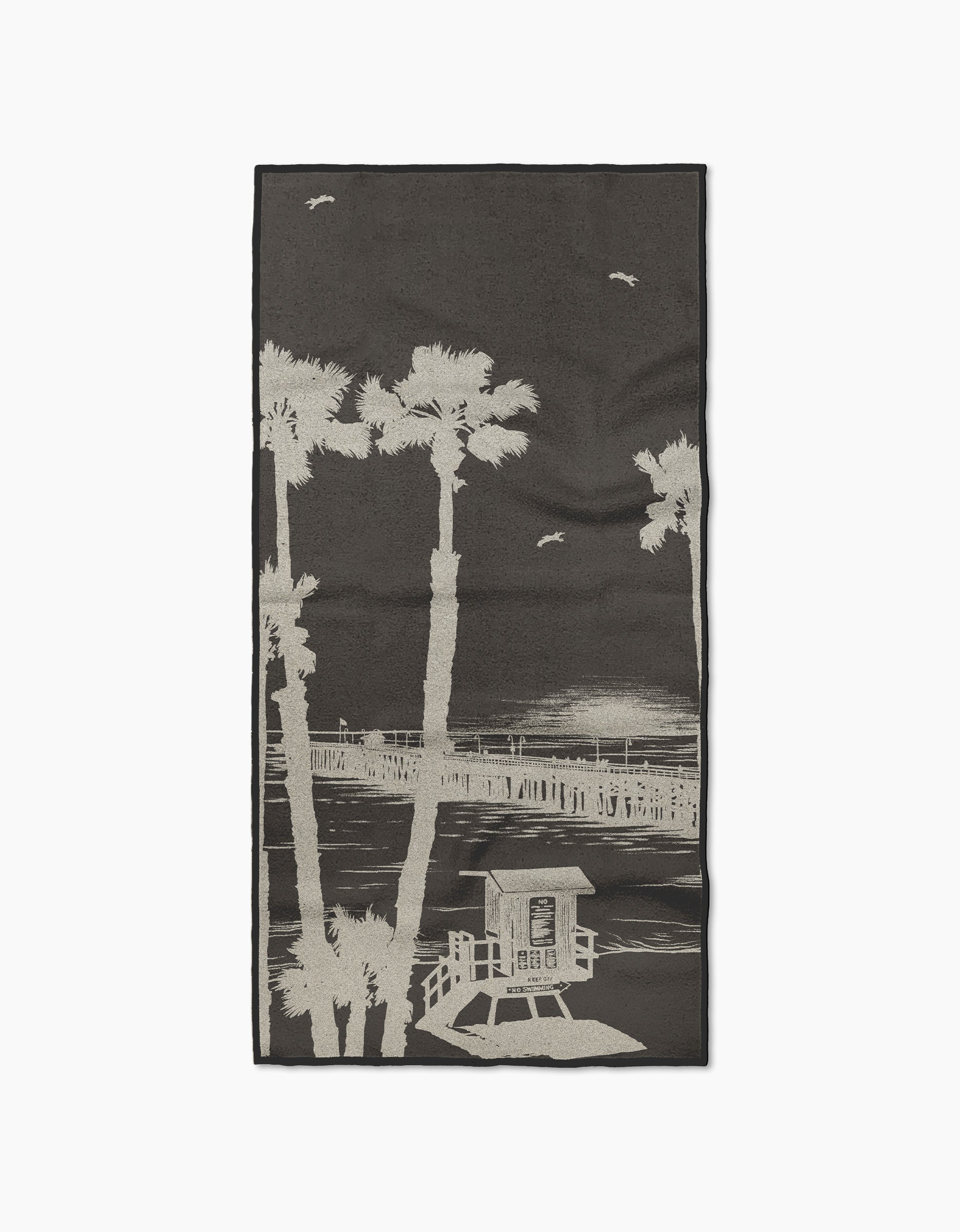 The Geometry Lost In Clemente beach towel features a monochrome palm tree, pier, and lifeguard tower design. Made from recycled materials, it’s fast drying and perfect for comfort at the beach.