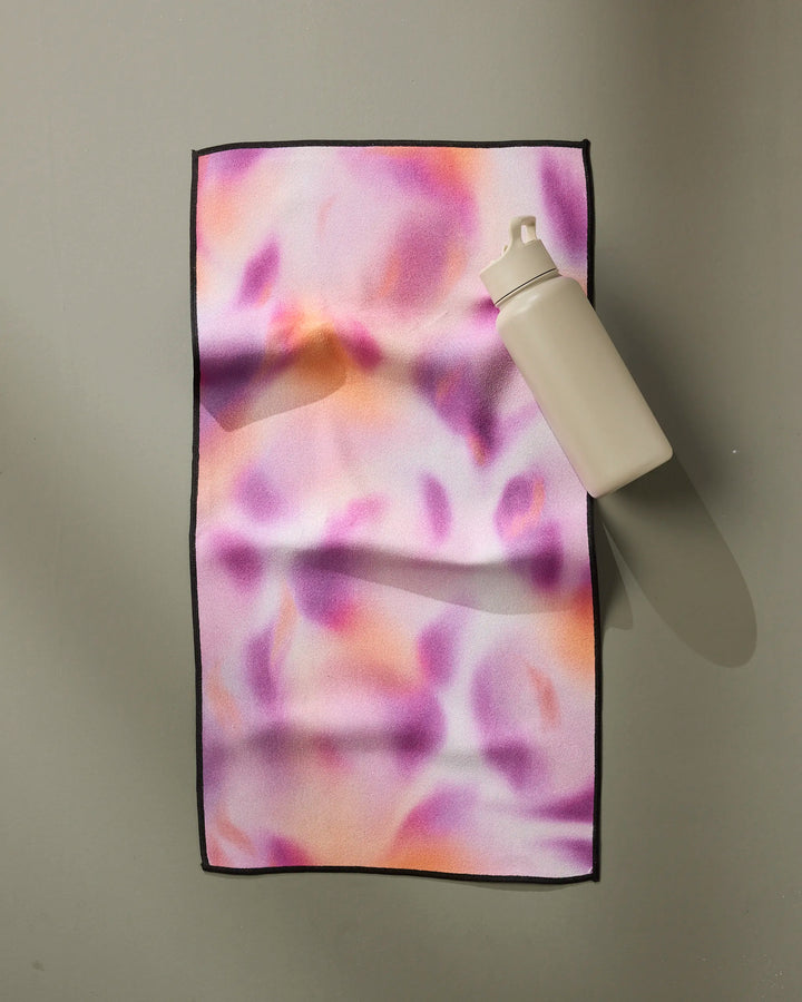A white water bottle sits atop the GEOMETRY Magenta fitness towel, showcasing a pink, purple, and orange abstract pattern on a smooth, neutral surface.