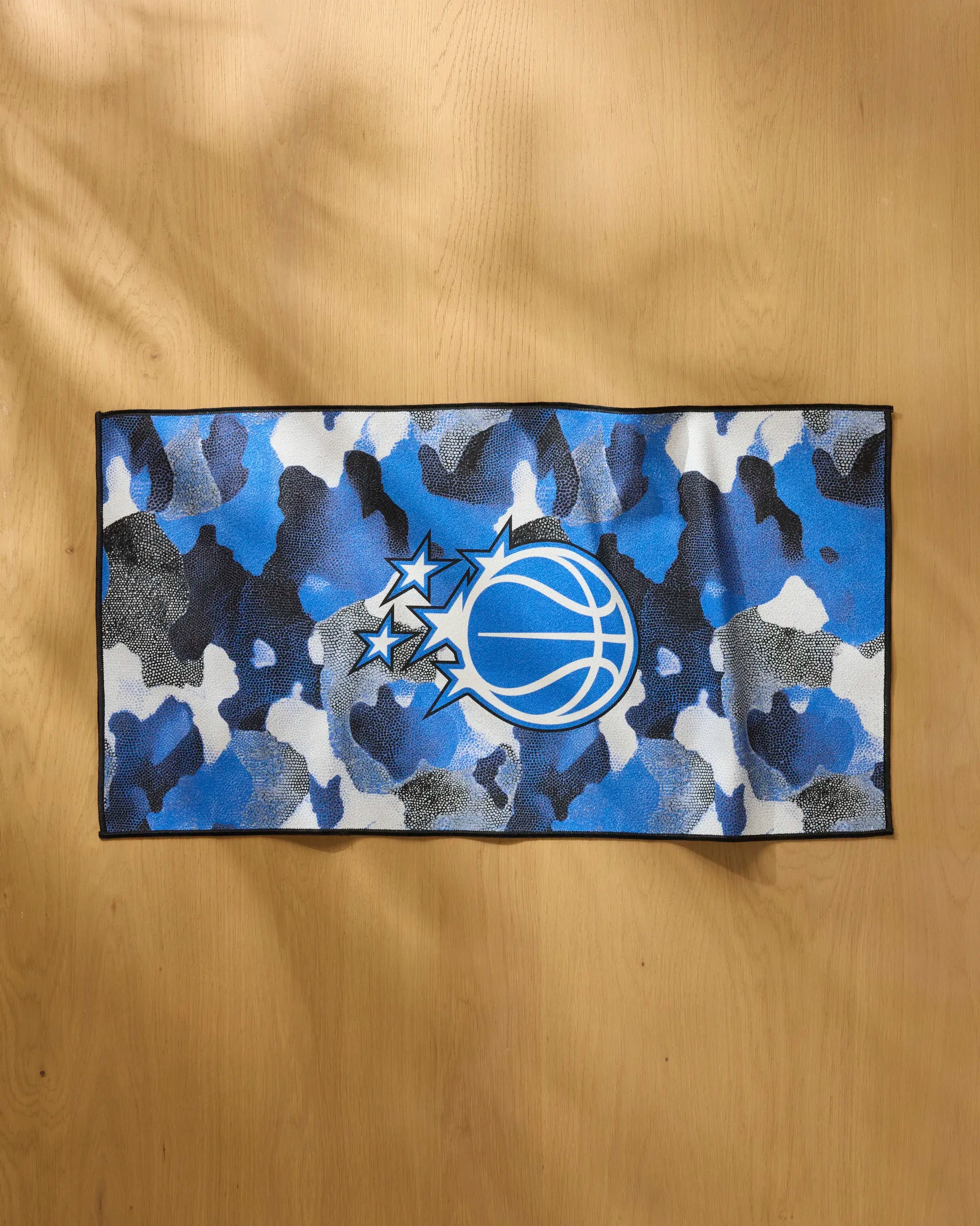 The Magic Camo towel by NBALAB x Geometry is a rectangular fitness towel with a blue, white, and black camo pattern and a basketball with stylized stars, shown on a wooden surface.