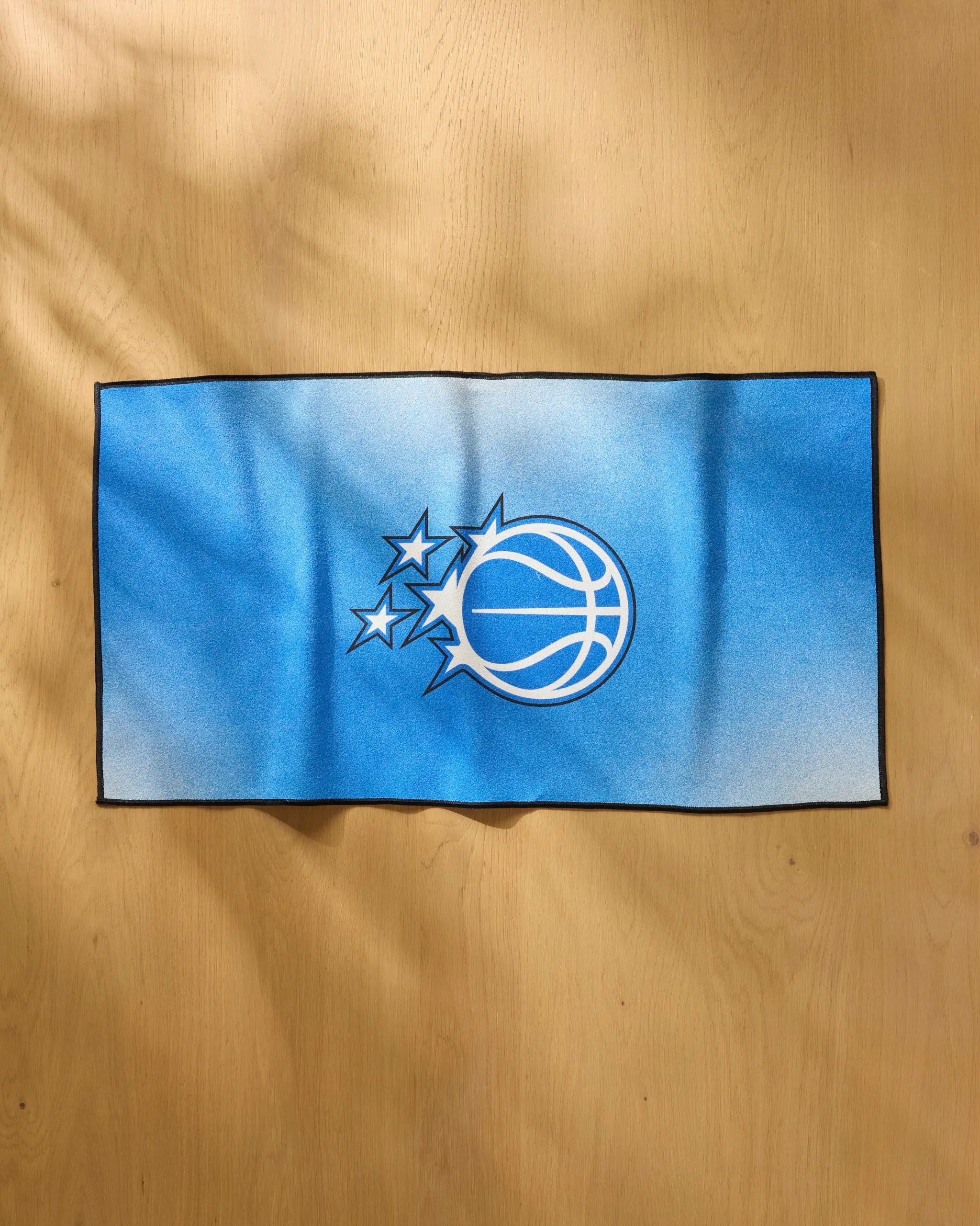 A blue rectangular "Magic Fade" fitness towel by NBALAB x Geometry features the Orlando Magic logo with three white stars and a black-and-white basketball on a light wooden surface.