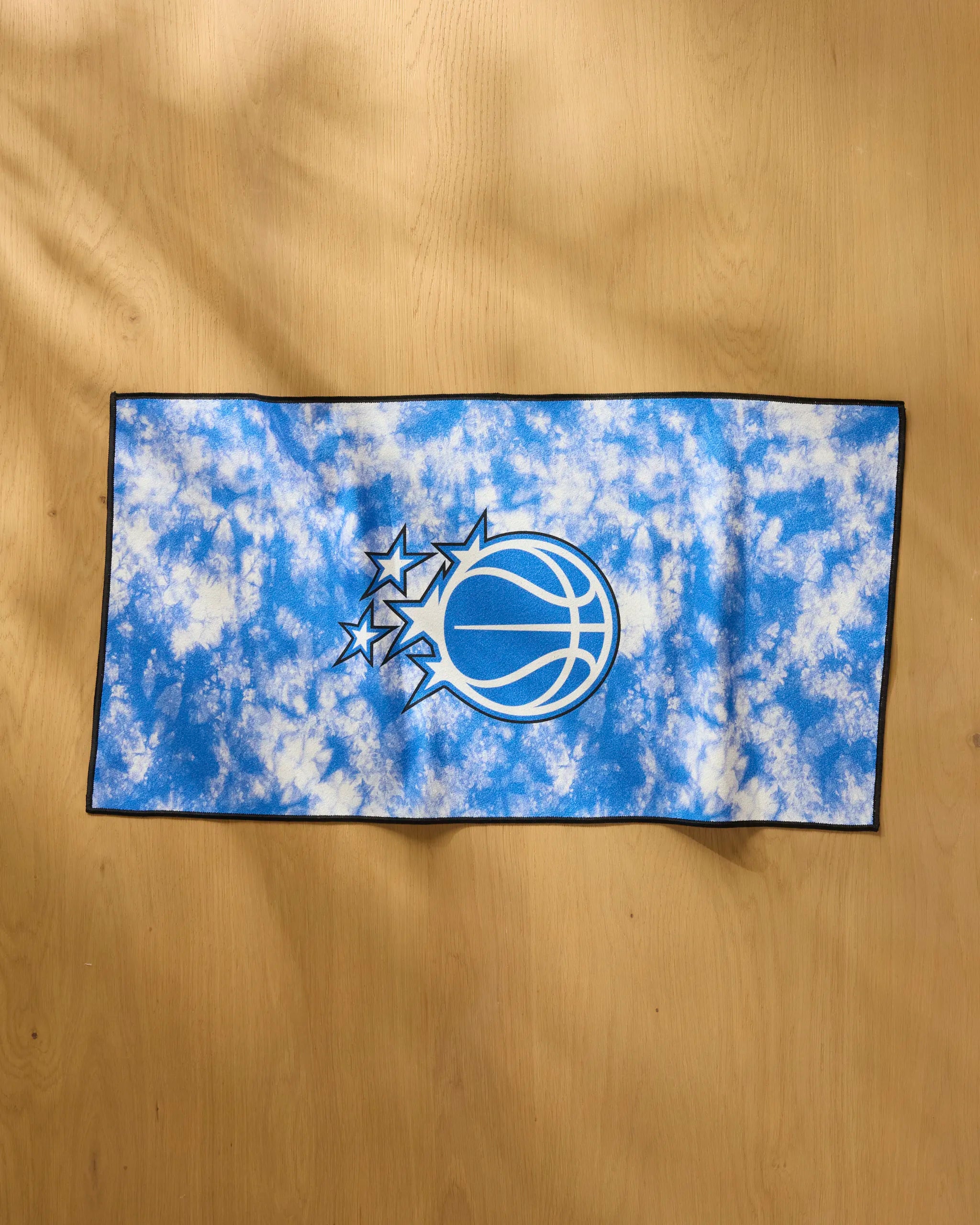 The Magic Flash by NBALAB x Geometry is a rectangular blue and white tie-dye NBA fitness towel with a black border and the Orlando Magic logo centered, shown on a light wooden surface.