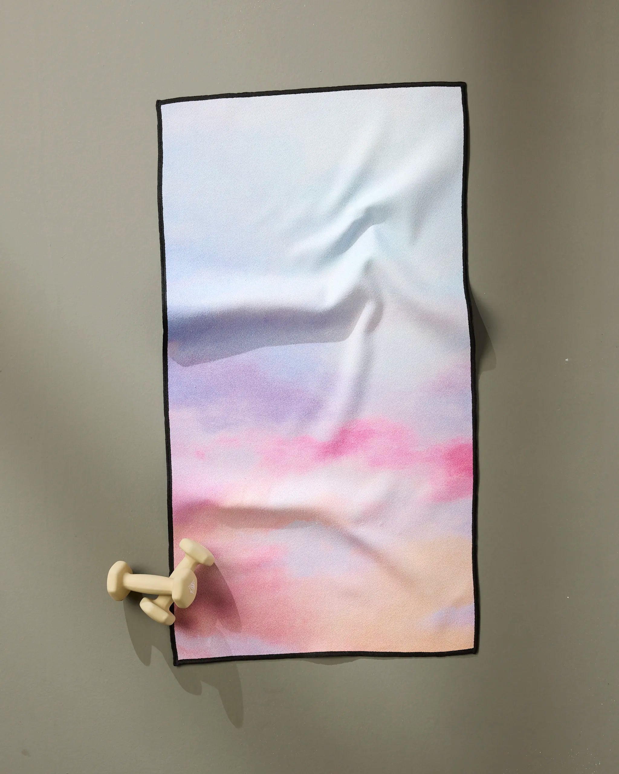 The "Magic In The Sky" fitness towel by Stefanie Bales, featuring pastel pink, blue, and beige hues, hangs on a gray wall. Ultra-absorbent and quick dry, it’s paired with two cream dumbbells at the bottom left corner.