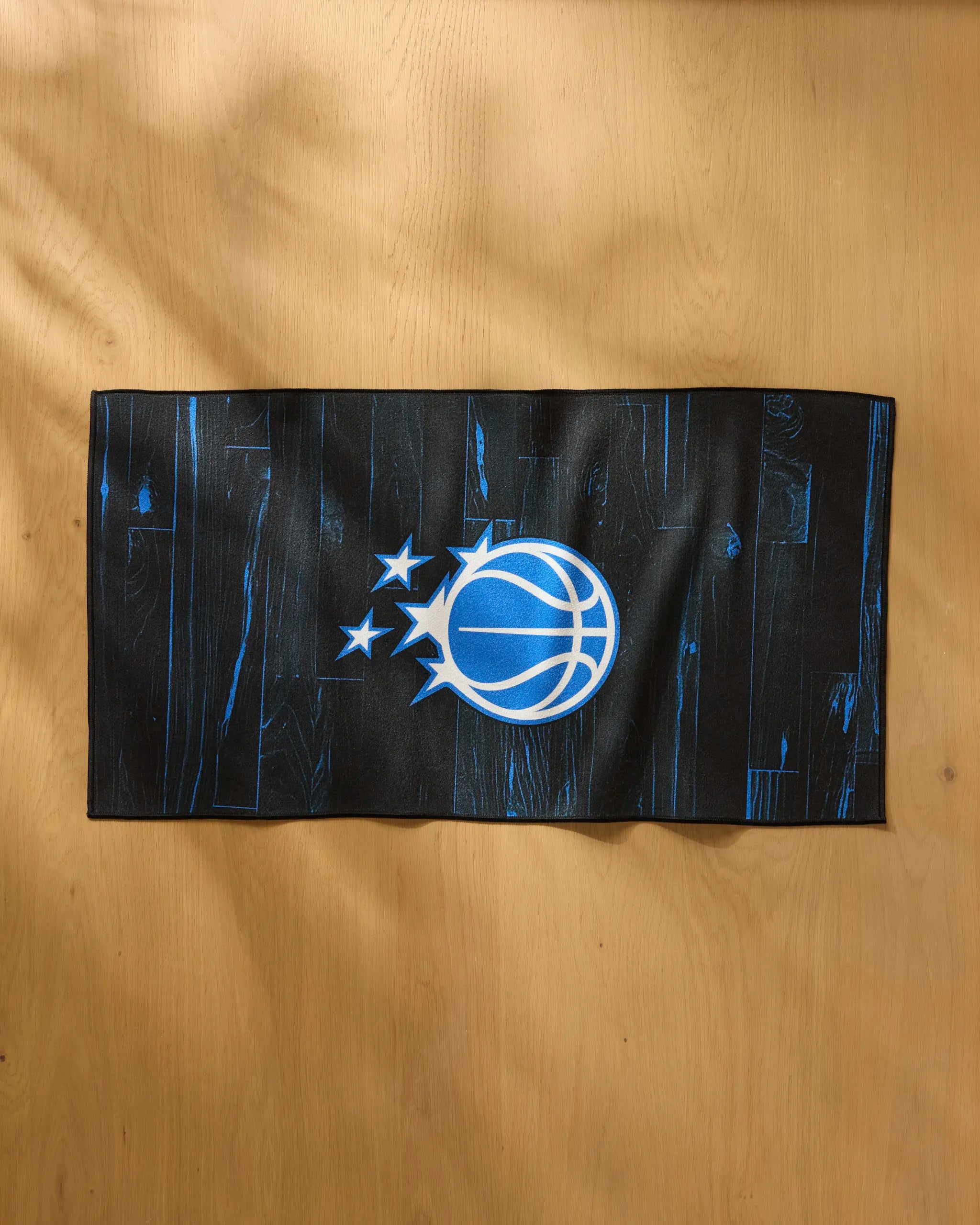 The Magic Zone fitness towel by NBALAB x Geometry, featuring blue wood-grain patterns and the Orlando Magic logo, is displayed on a light wooden surface.