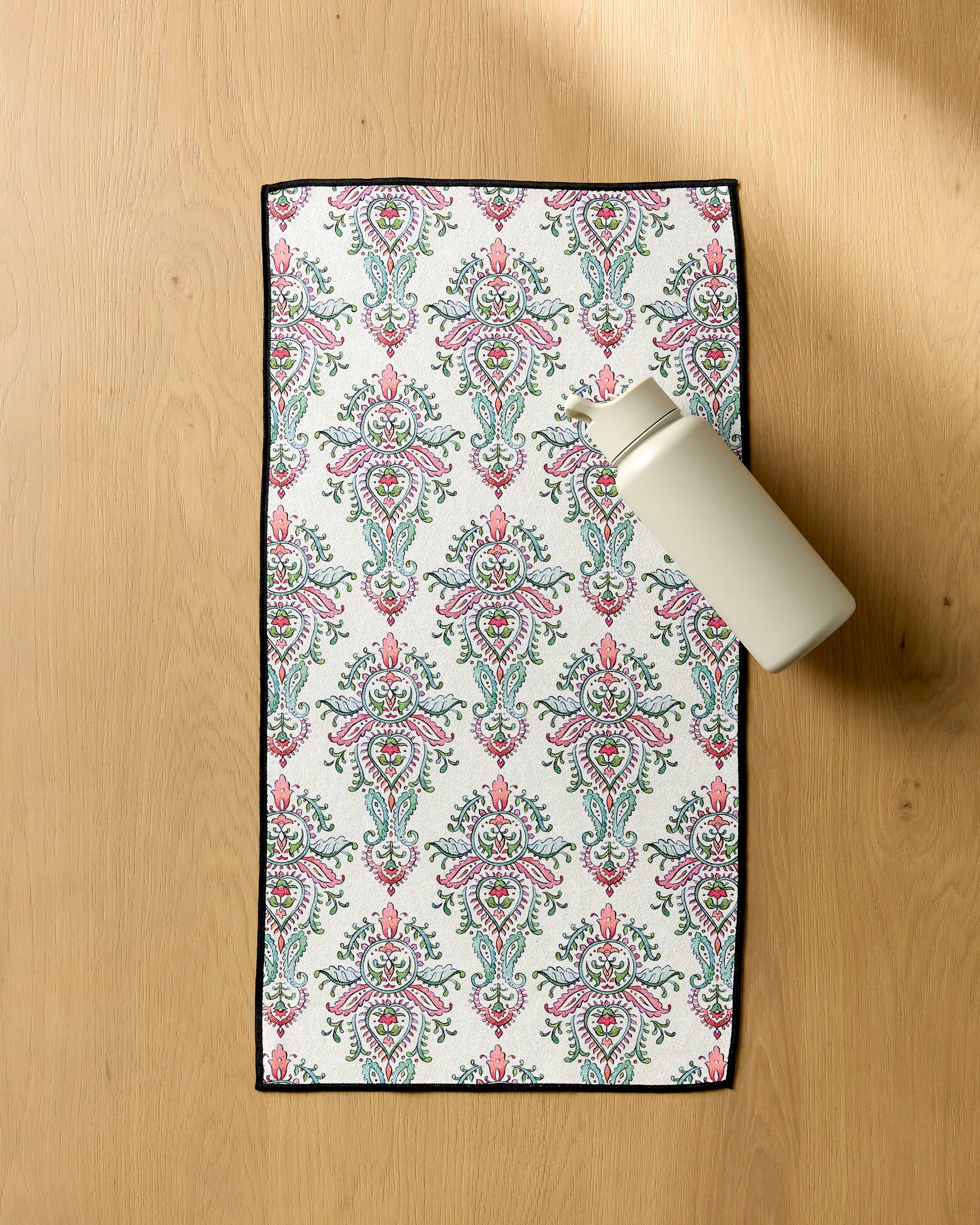 The Maayan Alper-Swan Memento—a white water bottle—rests on a rectangular, ultra-absorbent fitness towel with a pink and green ornate floral pattern, displayed on a light wooden surface.