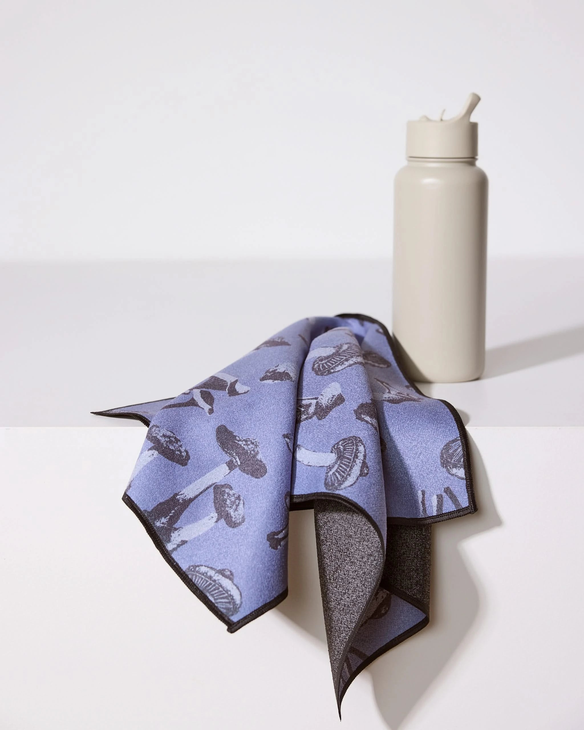 A gray water bottle with a straw sits on a white surface next to Geometry’s ultra-absorbent Moody Autumn Mushrooms fitness towel, draped in purple and adorned with a black mushroom pattern.