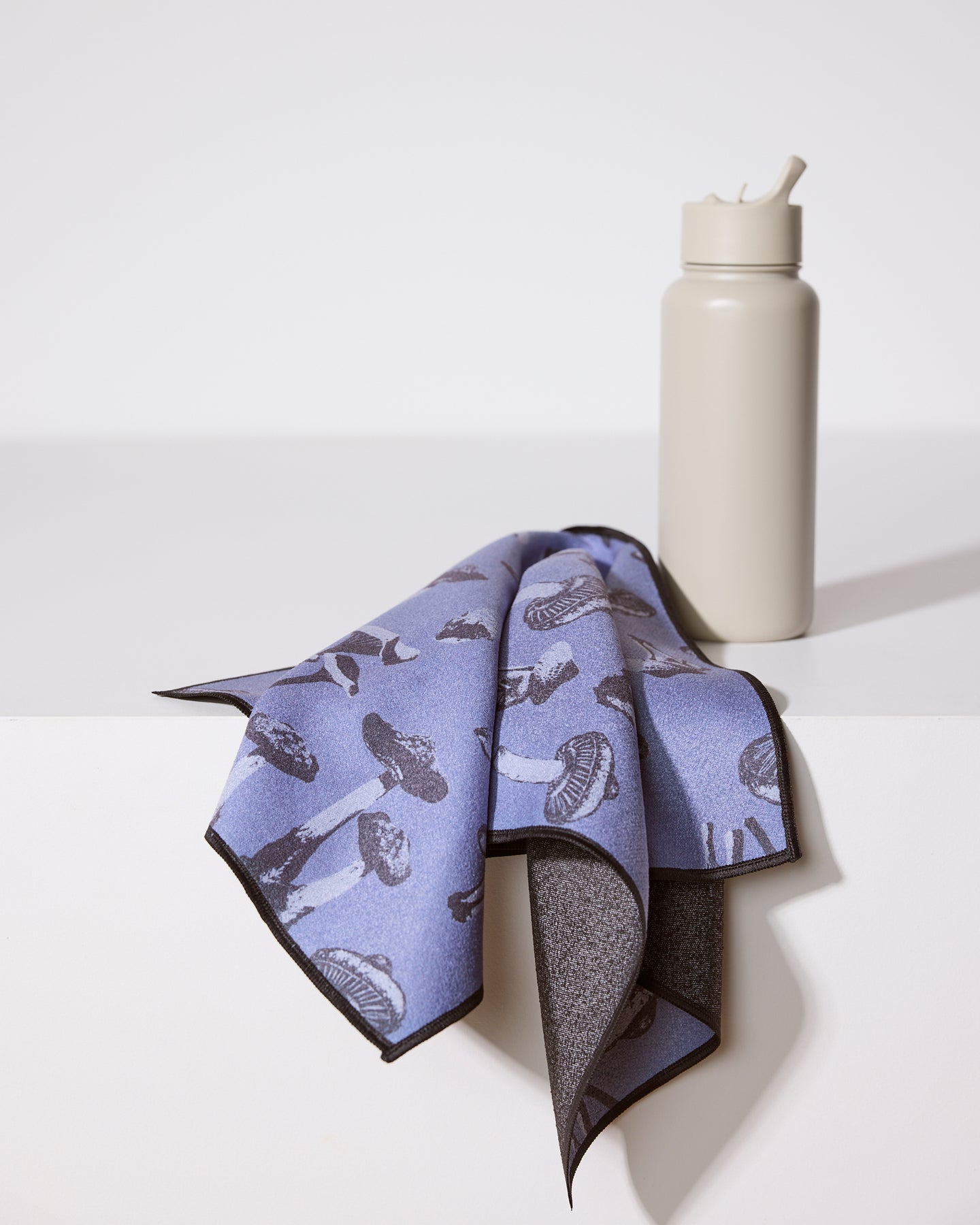 A gray water bottle with a straw sits on a white surface next to Geometry’s ultra-absorbent Moody Autumn Mushrooms fitness towel, draped in purple and adorned with a black mushroom pattern.