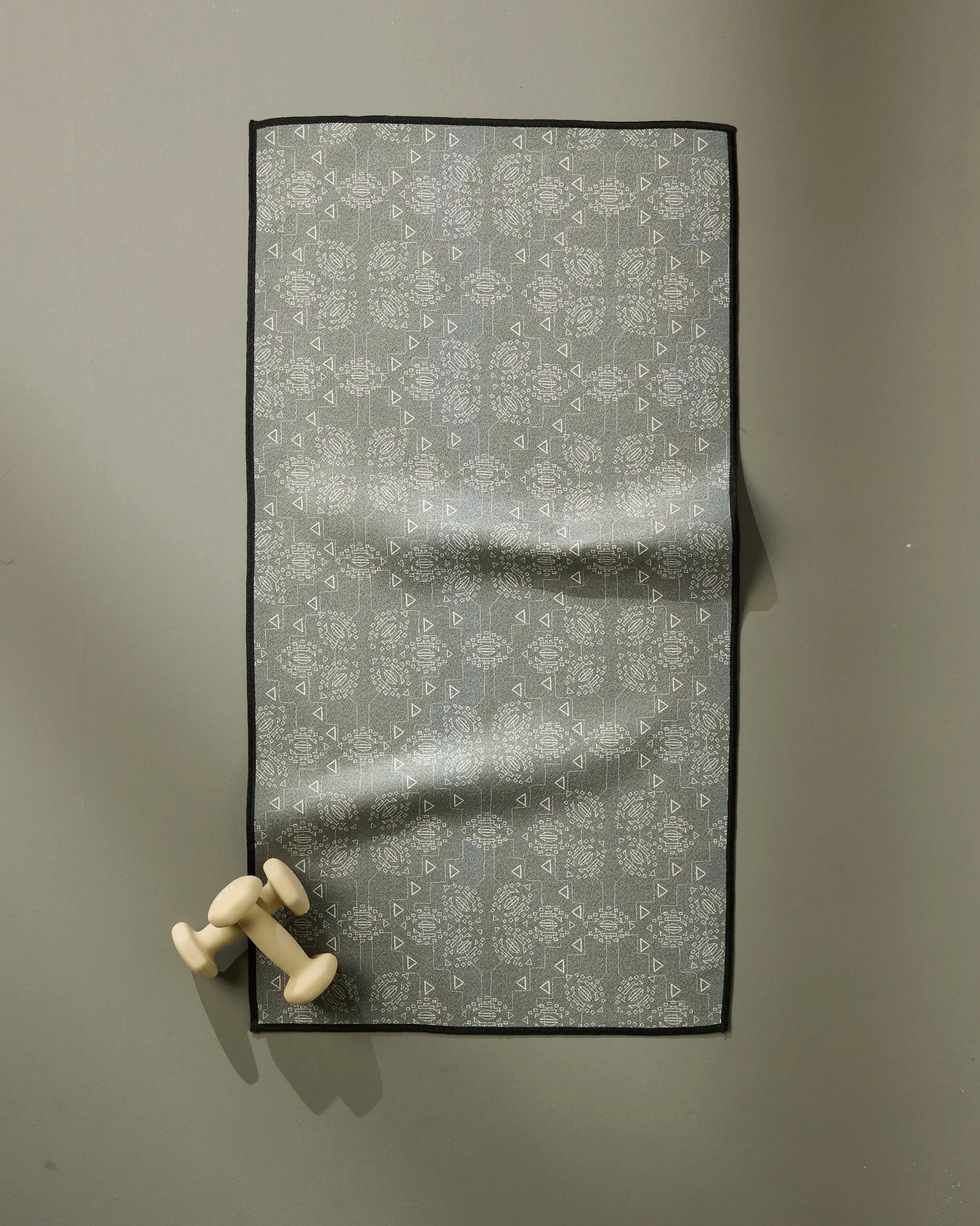 The Ash Cascade Mountain Bloom patterned grey fitness towel with a black border hangs on a wall, ultra-absorbent and fast drying, paired with small light-colored dumbbells at the bottom left corner.