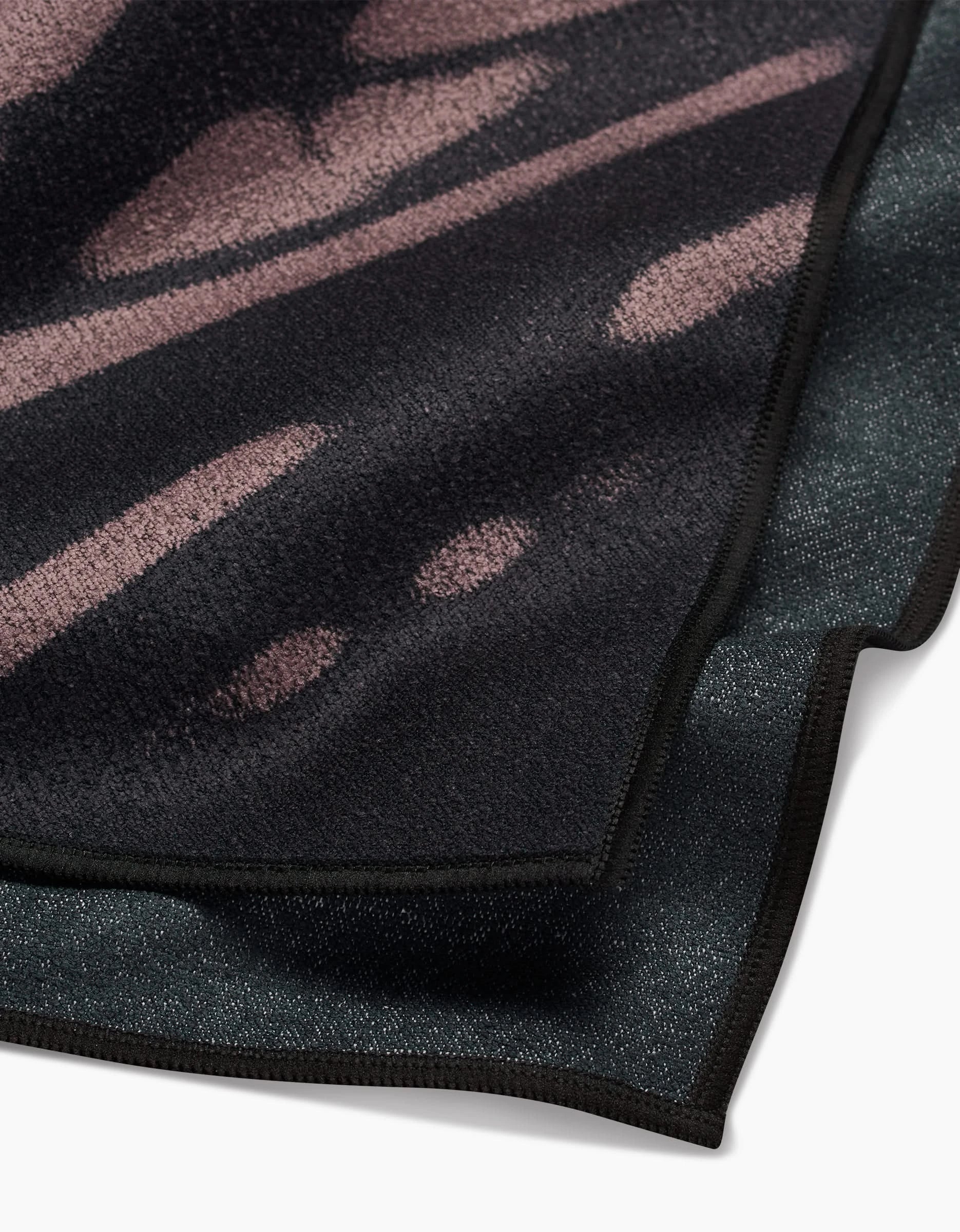 A close-up of two overlapping GEOMETRY blankets: Palm Shadow, made from ultra-absorbent Geoweave fabric with an abstract purple and black print, and a solid dark gray blanket, both featuring black stitched edges and a soft, textured surface.