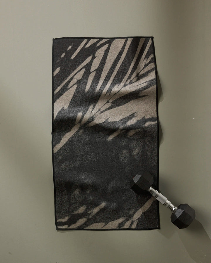 The GEOMETRY Palm Shadow ultra-absorbent fitness towel in a black and beige pattern is laid flat on a gray surface, with a black hexagonal dumbbell resting on its lower right corner.
