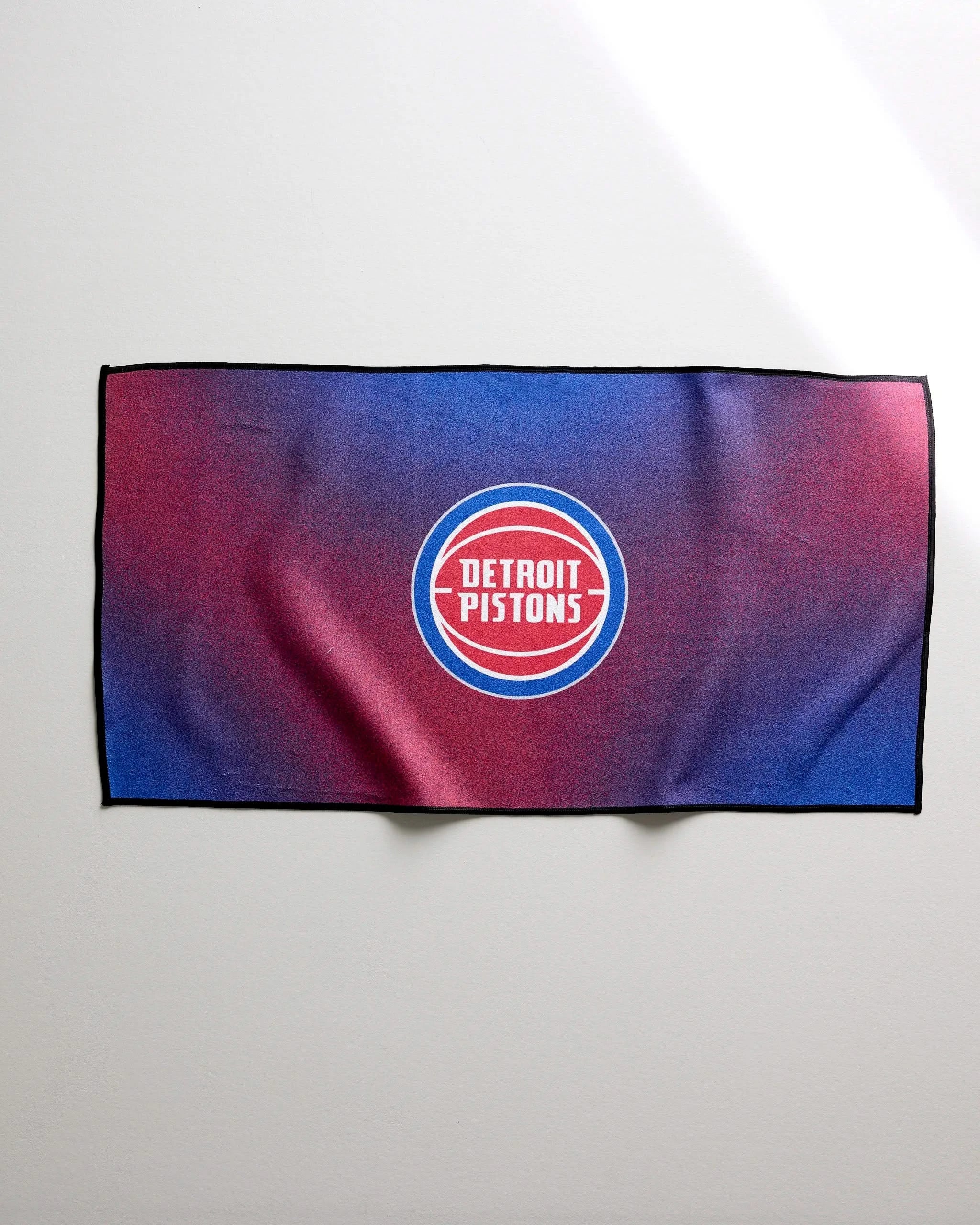 The NBALAB x Geometry Pistons Fade is a rectangular fitness towel with a central Detroit Pistons logo and a blue-to-red gradient, shown against a plain light wall.