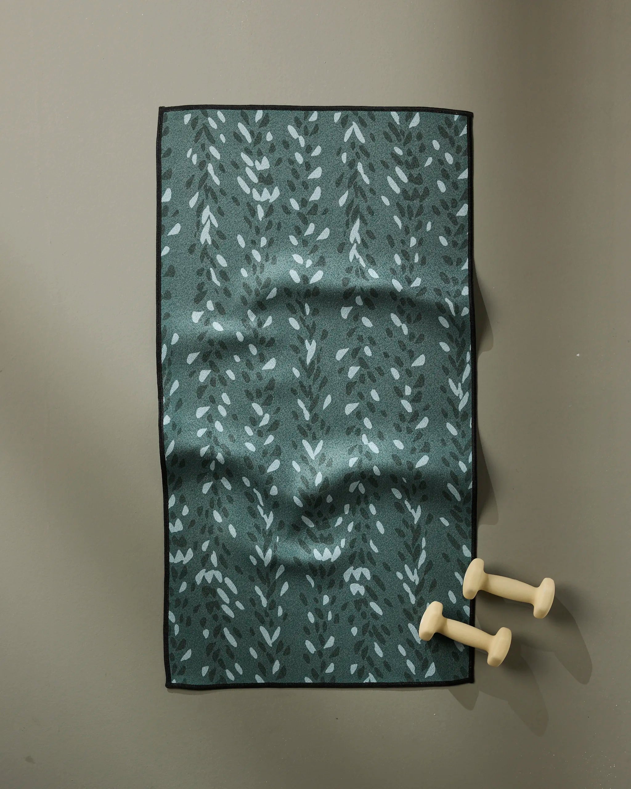 The GEOMETRY Pose mat, dark green with a light blue leafy design and made from recycled materials, lies flat on a gray surface. Two small beige hand weights are placed near its lower right corner.